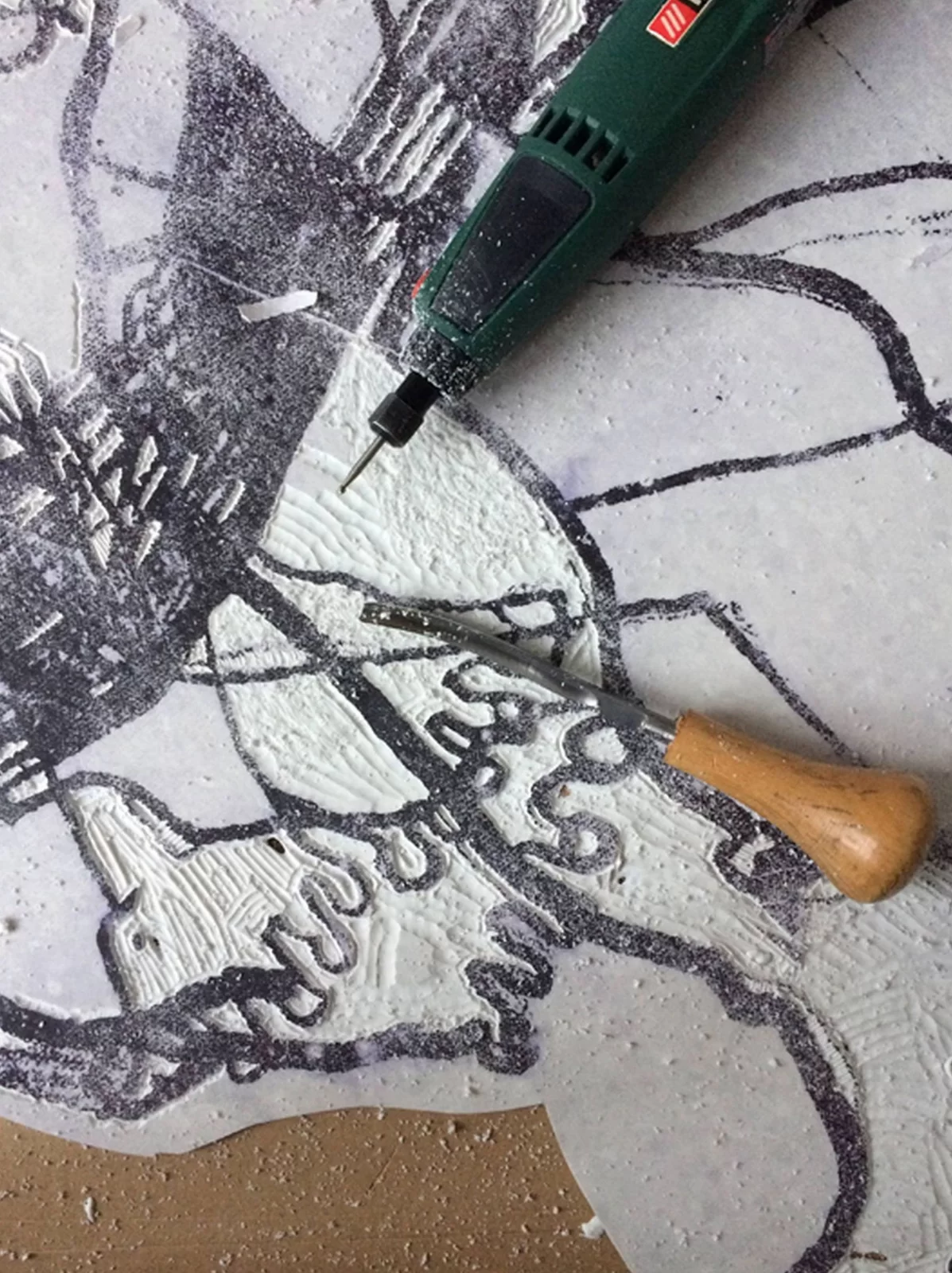 Ilustracja przedstawia narzędzia do grafiki. Na linorycie leży rylec, obsadzony w drewnianą rękojeść, a powyżej elektryczne dłutko. Na linoleum zostały już wycięte fragmenty. Sporządzony wcześniej czarny rysunek pozostał niewycięty.