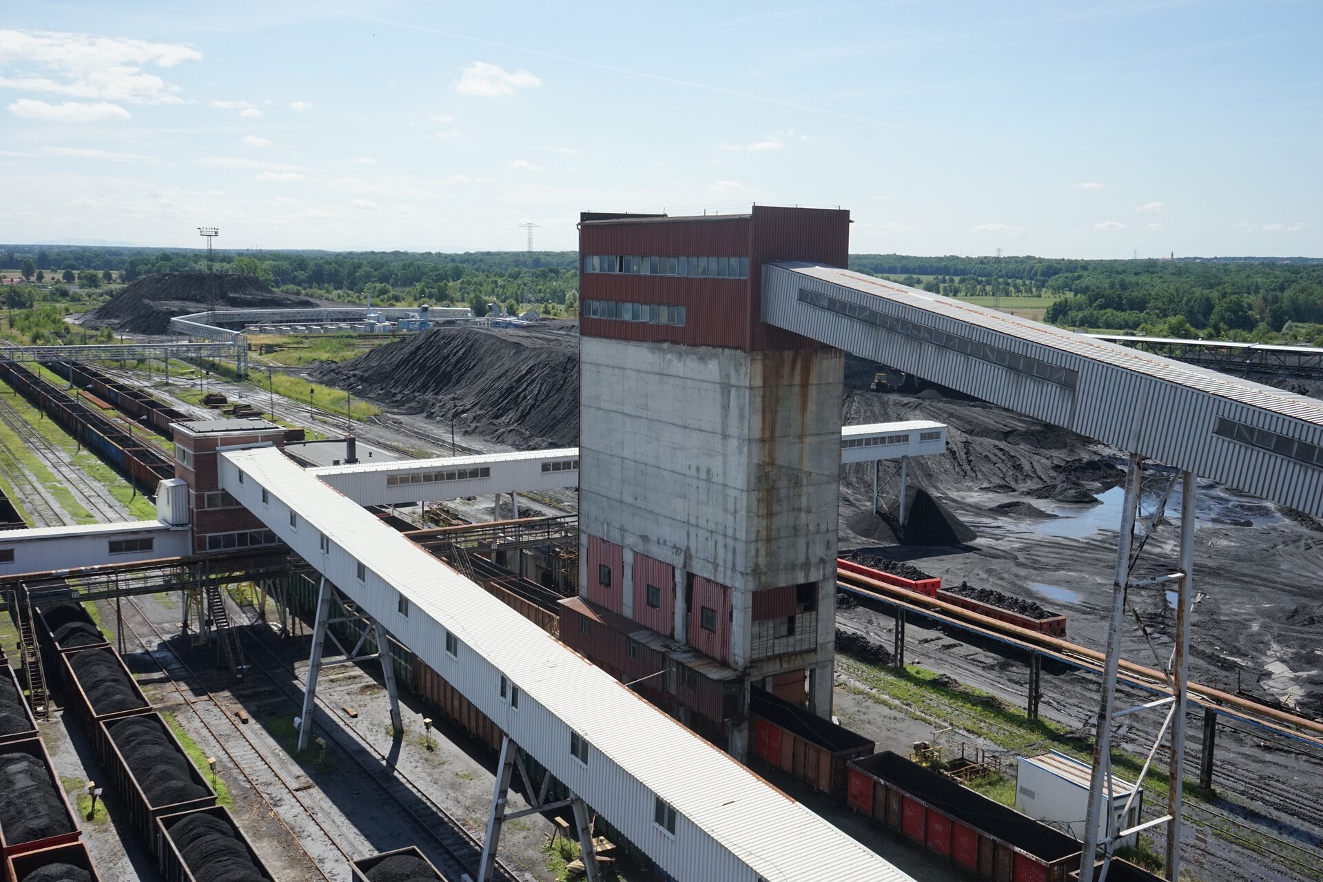 Na zdjęciu widoczny jest teren przemysłowy widoczny z góry. W centralnej części zdjęcia znajduje się duży budynek o kształcie prostopadłościanu. Do górnej części budynku dołączony jest most transportowy. Most pod postacią łącznika ma kształt prostopadłościanu. Most znajduje się na stalowych filarach. Łącznik wykonany jest z blachy falistej. Budynek znajduje się pomiędzy torami. Na torach stoją wagony kolejowe. Wagony są załadowane węglem. W tle zdjęcia widać hałdy z węglem. Hałdy mają kształt piramidy.Poniżej zdjęcia widoczny jest przycisk multimedialny z nagraniem audio. Treść nagrania jest zgodna z tytułem zdjęcia.
