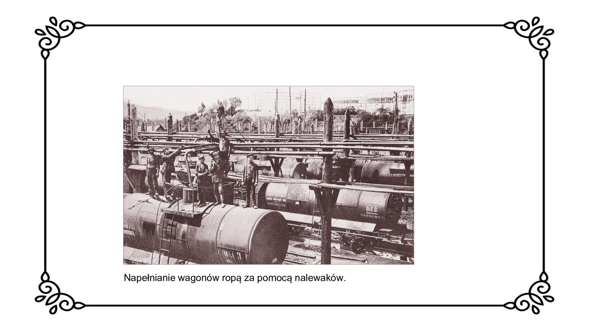 Slajd przedstawia proces napełniania cystern kolejowych ropą za pomocą nalewaków wprost z rurociągu, biegnącego od kopalni na stację kolejową. Widoczne są poustawiane na torach kolejowych, w rzędzie cysterny. Na górze jednej z nich stoją mężczyźni i ją napełniają. 