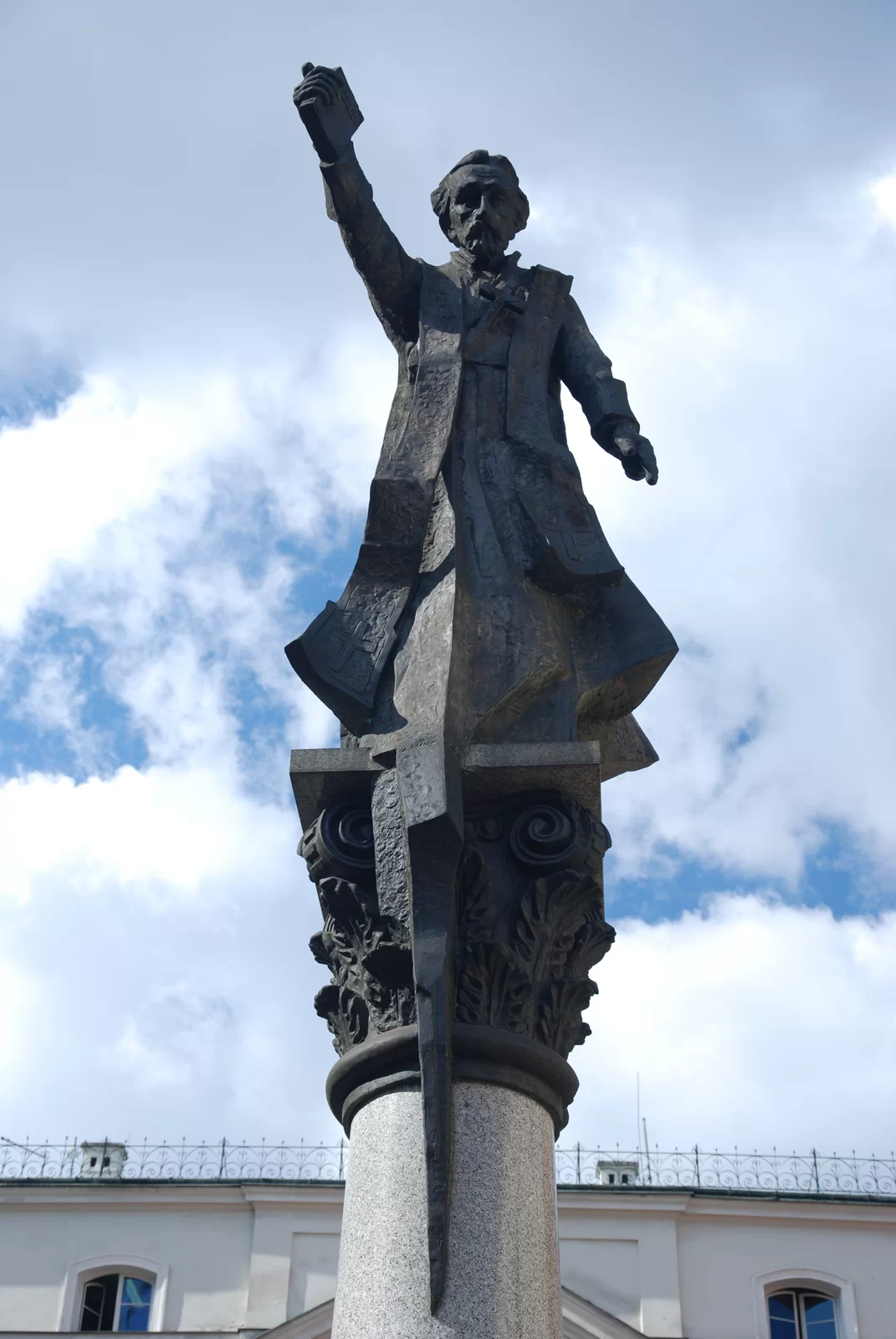 Zdjęcie przedstawia pomnik. Na owalnej kolumnie znajduje się postać stojącego mężczyzny. Jest w długiej szacie, ma przewieszoną przez szyję stułę. Trzyma uniesioną w górę prawą rękę, a w dłoni książkę.