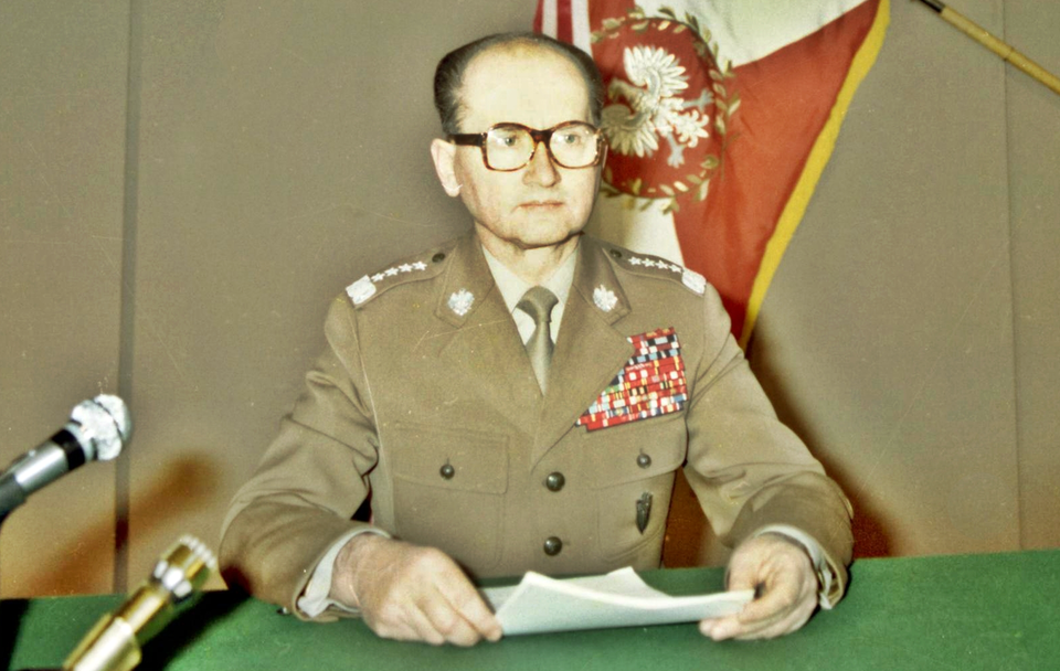 Zdjęcie przedstawia dojrzałego, łysiejącego mężczyznę w mundurze generalskim i okularach w grubych oprawkach. Mężczyzna siedzi, za blatem przykrytym zielonym suknem, na którym stoją mikrofony. Trzyma w dłoniach plik kartek.