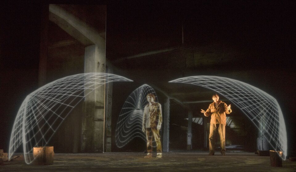 Fotografia przedstawia przykład zastosowania technologii mapowania w projekcji scenicznej bohatera i wprowadzeniu efektów specjalnych, „Dedal – Ikar” autorstwa Lemieux Pilon 4D Art. Na pierwszym planie znajduje się postać w brązowym ubraniu. Na nią nałożona jest biała siatka. Z lewej strony ilustracji znajduje się niski pieniek drzewa, nad którym jest biała siatka. Z prawej strony ilustracji również znajduje się pieniek drzewa, nad którym jest biała siatka. Pod tą siatką stoi także mężczyzna w ubraniu w odcieniach brązu. Jego ręce są podniesione na wysokości łokci. Mężczyźni są w wysokim, pustym pomieszczeniu. Tło tej fotografii jest ciemne.