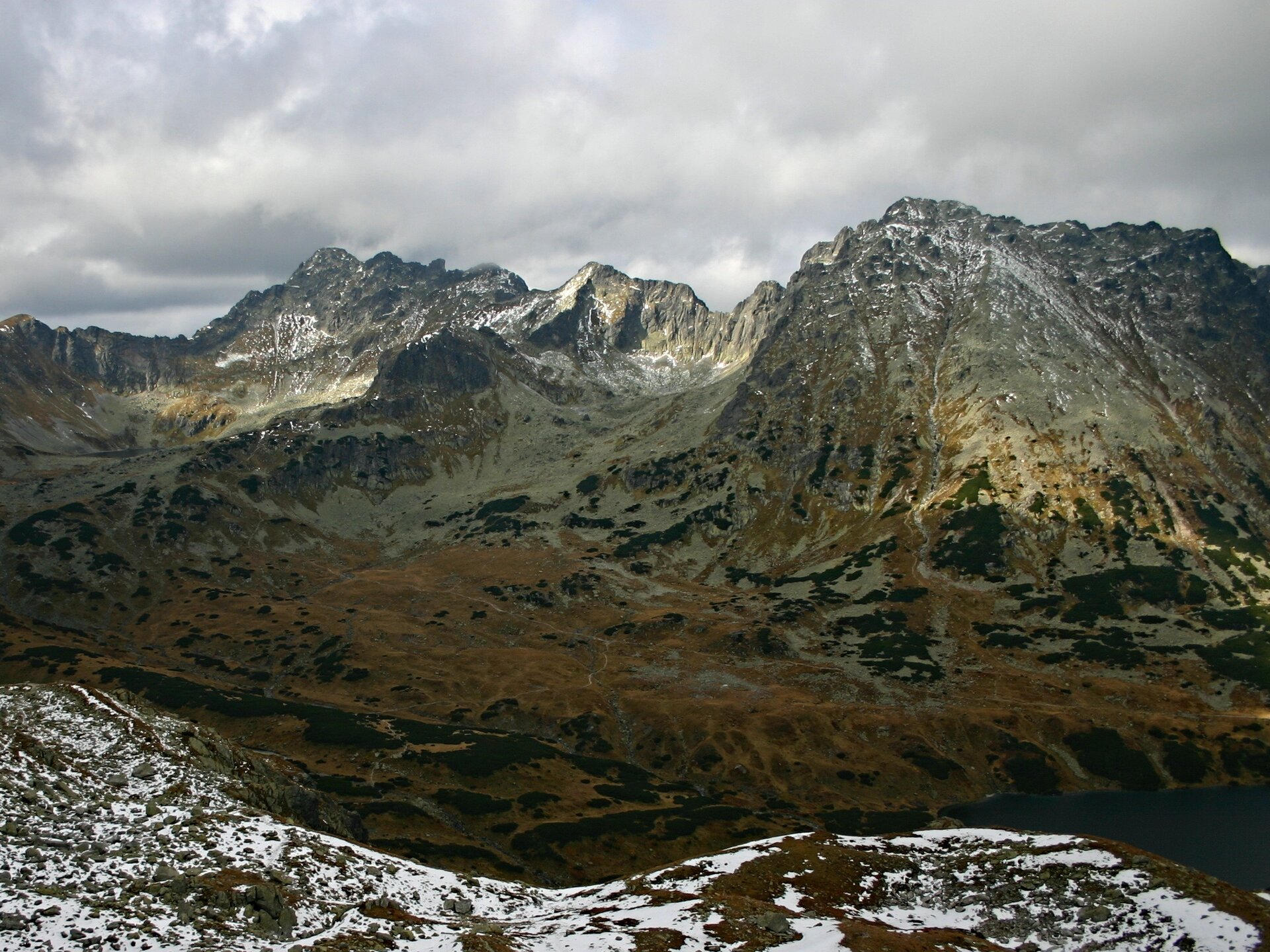 Na fotografii widoczne jest szare, z ostrymi szczytami pasmo górskie, porośnięte pomarańczową trawą. U podnóża gór widoczna jest rozległa przełęcz, pokryta w połowie śniegiem. W tle widoczne są szare chmury.