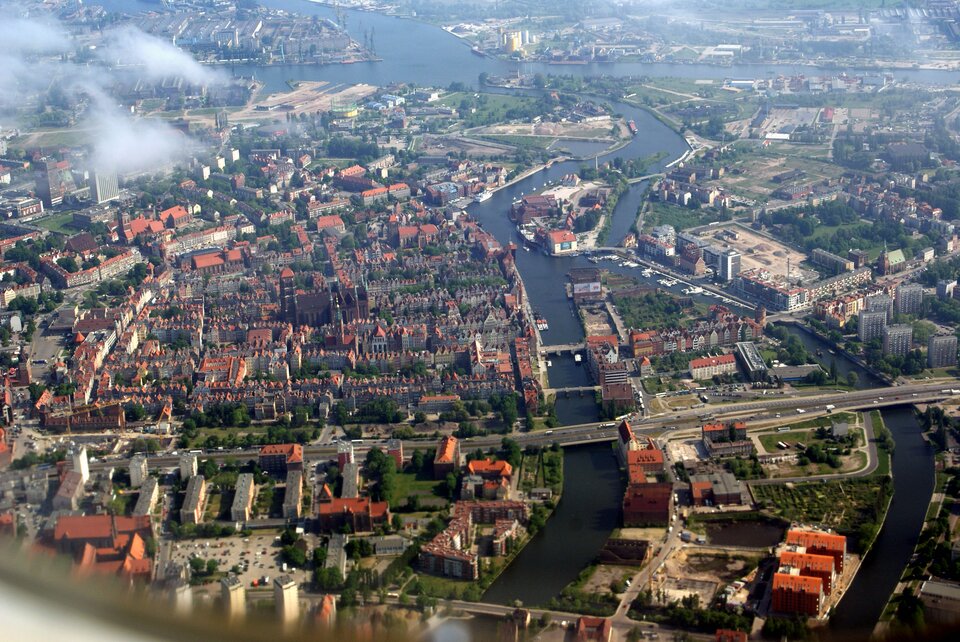 Na fotografii widać fragment Gdańska ujęty z lotu ptaka. Charakteryzuje się on niską, gęstą, miejską zabudową. Widoczne jest również Stare Miasto z górującym nad nim ceglanym Kościołem Mariackim. Miasto przecina rzeka Motława. Są na niej liczne mosty oraz wyspy. 