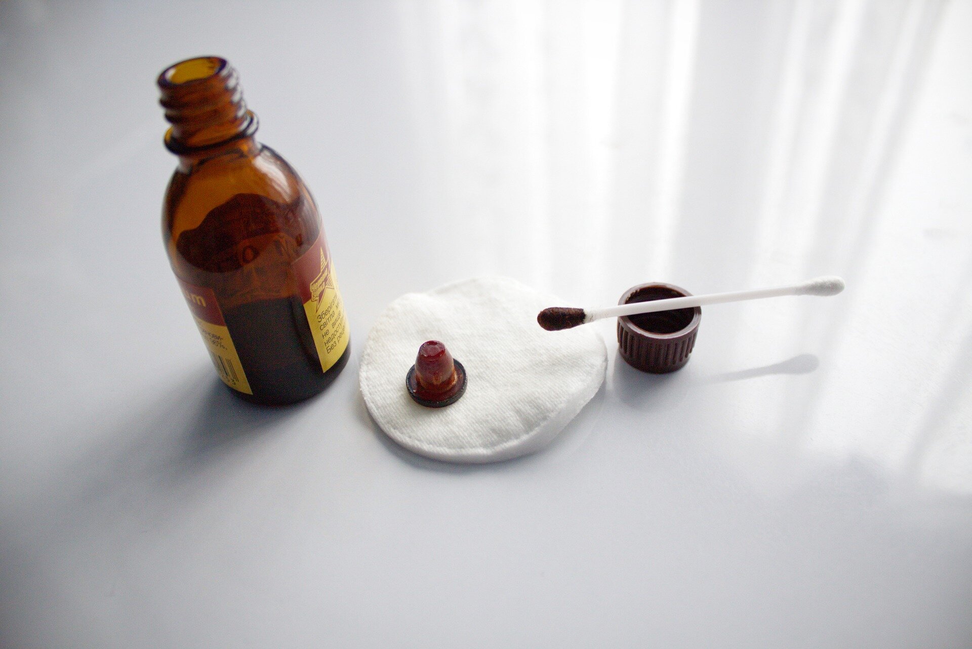 Ilustracja przedstawia szklaną, ciemną butelkę. Obok znajdują się bawełniany wacik i patyczek pokryte ciemną substancją.