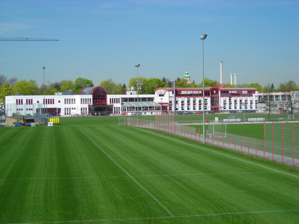 Zdjęcie przedstawia zieloną murawę boiska sportowego. Zostało one wykonane w pogodny dzień. Nieopodal boiska znajdują się niewysokie, charakterystyczne zabudowania. Trwają prace budowlane. Uchwycono fragment dźwigu. Trawa ma soczyście zielony kolor, jest niska. Murawę podzielono na prostokątne pola za pomocą białej farby.