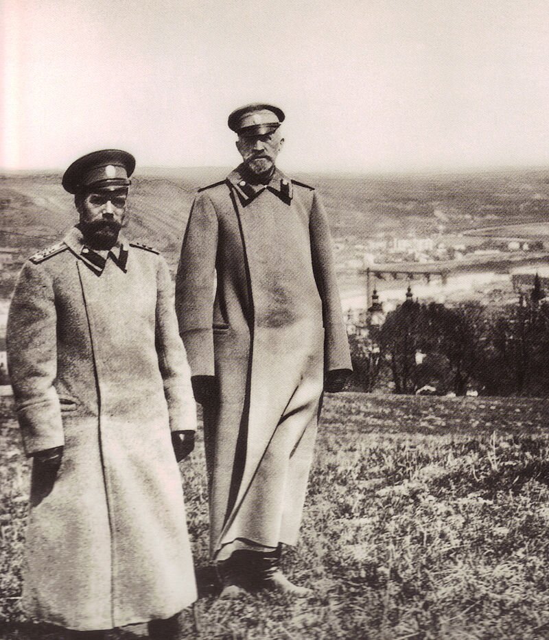 Na zdjęciu dwoje mężczyzn w płaszczach mundurowych. Na ramionach pagony. Czapki z herbem. Oboje mają dłuższe wąsy. Stoją na wzgórzu. W dolinie rozpościera się widok miasta. 