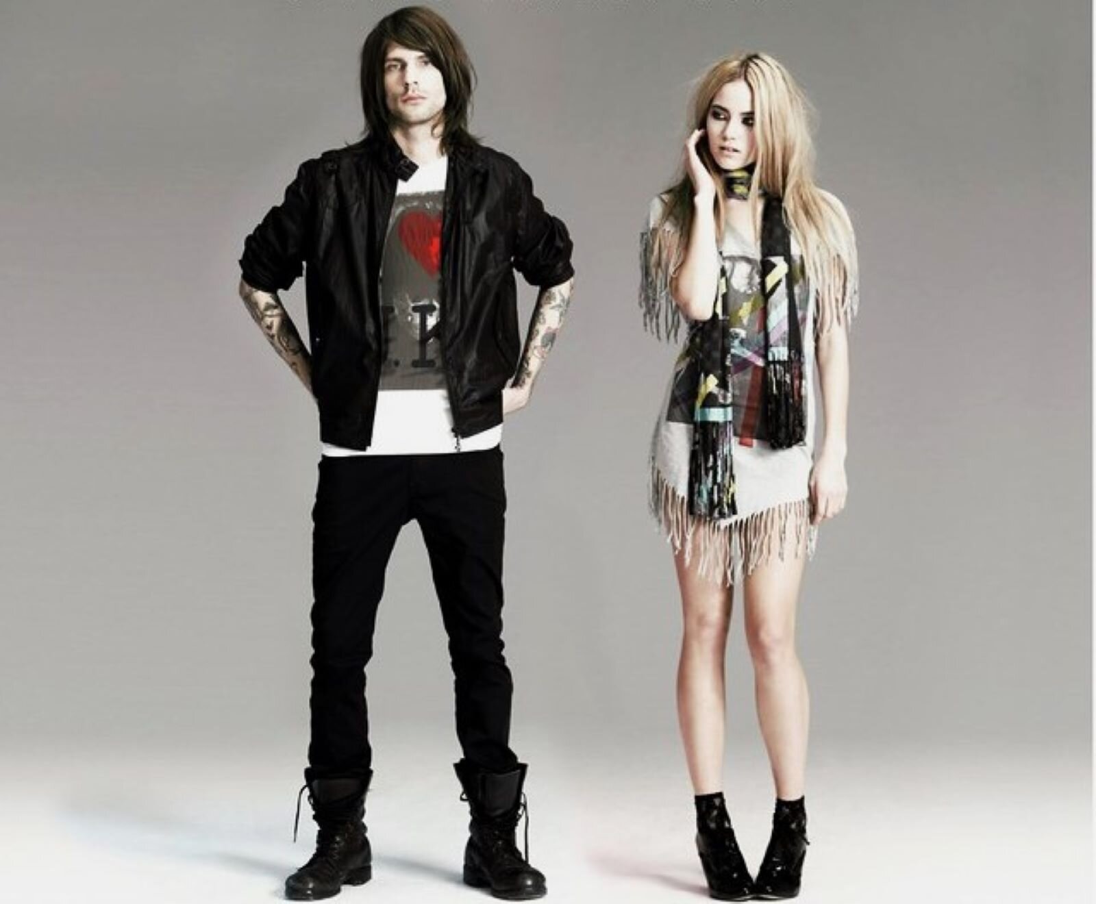 Zdjęcie przedstawia chłopaka i dziewczynę reklamujących ubrania. Chłopak jest ubrany w skórzaną kurtkę, długie spodnie i ciężkie buty za kostkę, stoi w wyzywającej pozie w rozkroku z wytatuowanymi rękami na biodrach. Dziewczyna obok jest krucha i delikatna, patrzy w bok, trzymając jedną rękę przy twarzy. Ma długie włosy, a na sobie krótką sukienkę z frędzlami na dole i ramionach,  szalik na szyi, a na gołych nogach buty za kostkę na obcasie.