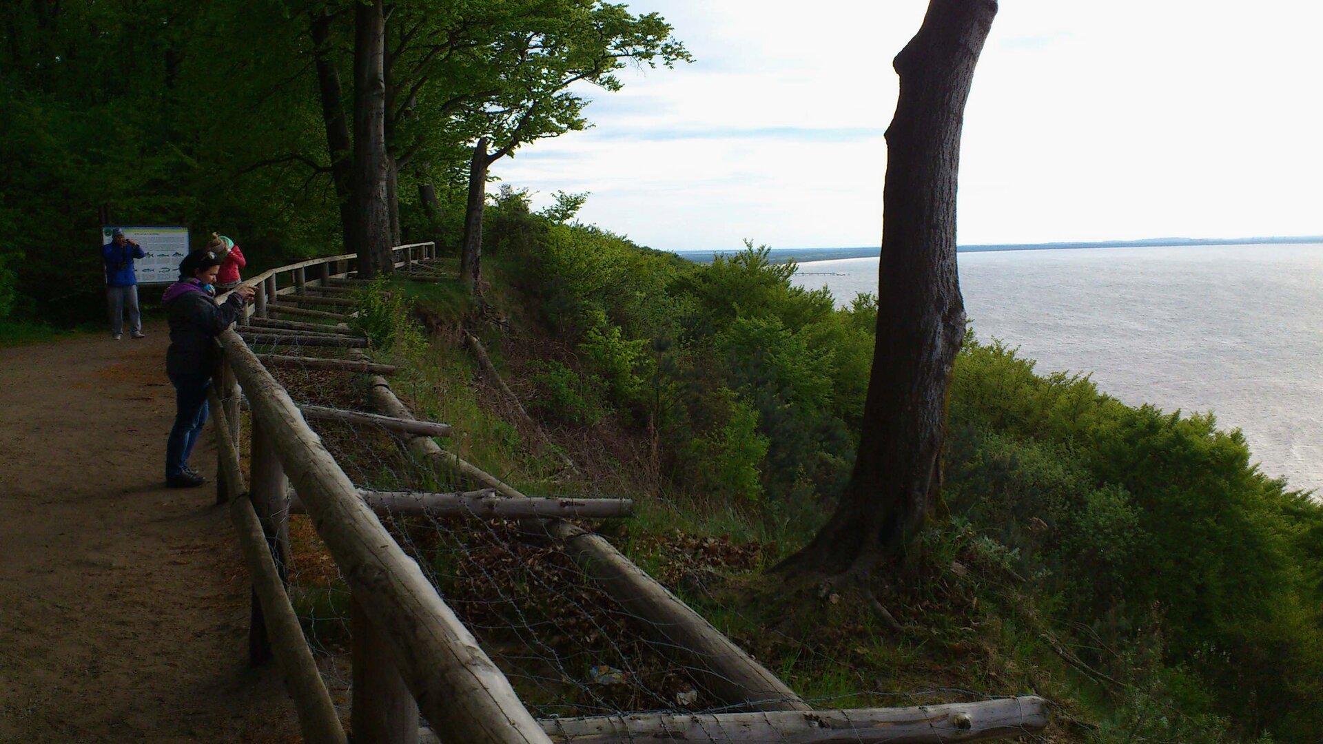 Na zdjęciu punkt widokowy ogrodzony drewnianą barierką, tablica informacyjna, w dole morze.