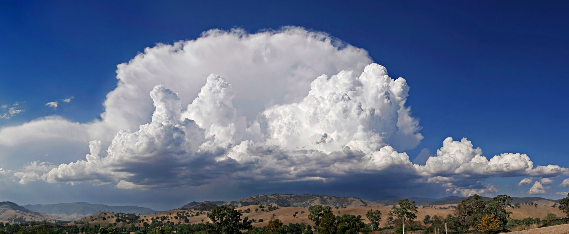 Ilustracja przedstawia chmury kłębiasto-deszczowe - cumulonimbusy. Są to chmury kłębiaste deszczowe – gęste chmury rozbudowane pionowo na wysokość kilku lub kilkunastu kilometrów. Na zdjęciu chmury te unoszą się nad pagórkami i niskimi górami. 