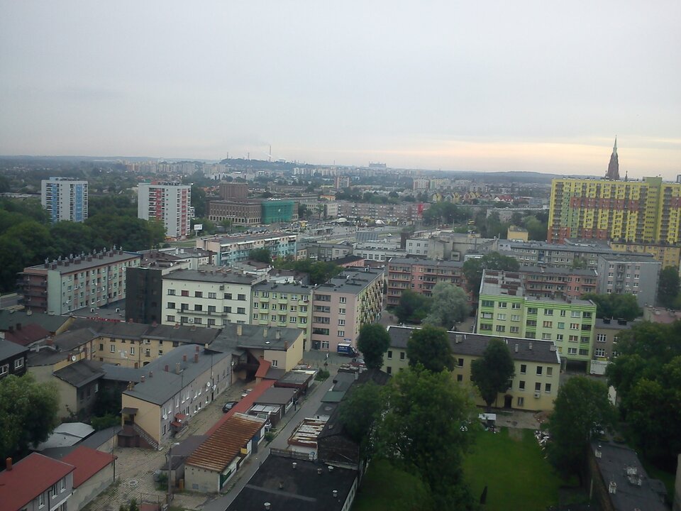 Na zdjęciu lotniczym zabudowa mieszkaniowa złożona z dość niskich budynków w kolorach zielonym, jasnozielonym, szarym. Część z nich ma ciemne dachy, a część czerwone. Pomiędzy budynkami po prawej stronie kadru drzewa, krzewy i trawniki. Obok budynków ulice. Na drugim planie po prawej stronie dziesięciopiętrowy szeroki wieżowiec w kolorze żółto‑zielono‑czerwonym. Zza niego wystaje wieża – najprawdopodobniej kościoła w znacznie starszej architekturze. W tle zabudowania przemysłowe i pochmurne niebo, które zajmuje jedną trzecią kadru.