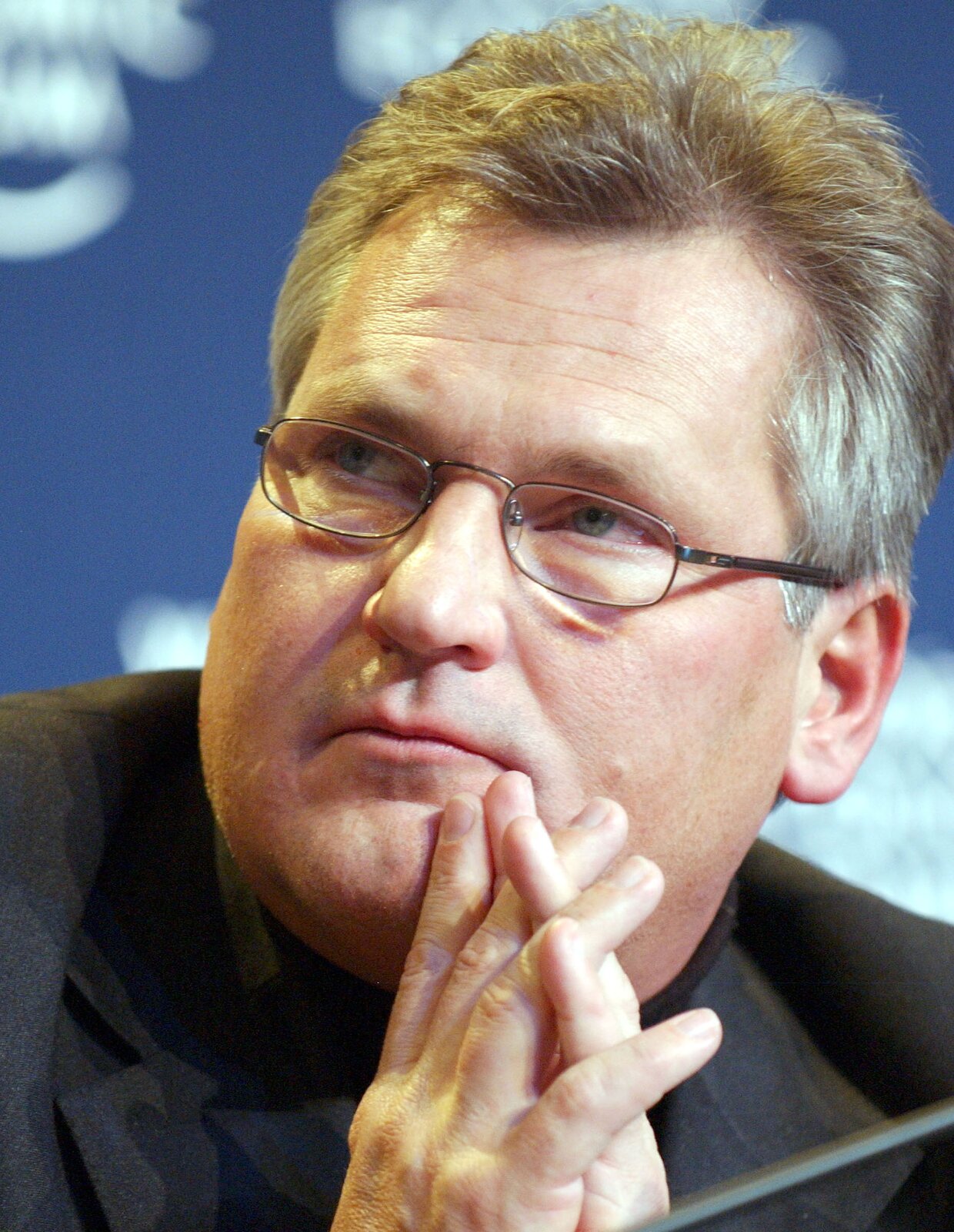 Fotografia portretowa przedstawiająca prezydenta Aleksandra Kwaśniewskiego. Mężczyzna w średnim wieku, twarz owalna, włosy szpakowate, zaczesane do góry. Okulary w drucianej oprawie. Mężczyzna ma przy twarzy złożone ręce.