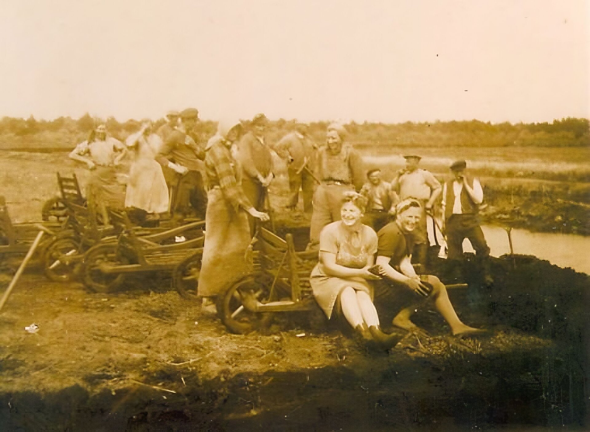 Zdjęcie przedstawia ludzi na polu przy drewnianych wózkach. Z jednej strony znajduje się koło, a z drugiej - drążki do pchania lub ciągnięcia wózka. Dwie kobiety siedzą na wózku z przodu, pozostałe osoby stoją między wózkami i przy nich. Są uśmiechnięci. Wśród nich znajdują się kobiety i mężczyźni. Kobiety ubrane są w spódnice, na głowie noszą chusty. Mężczyźni noszą kamizelki, na głowie mają czapki.