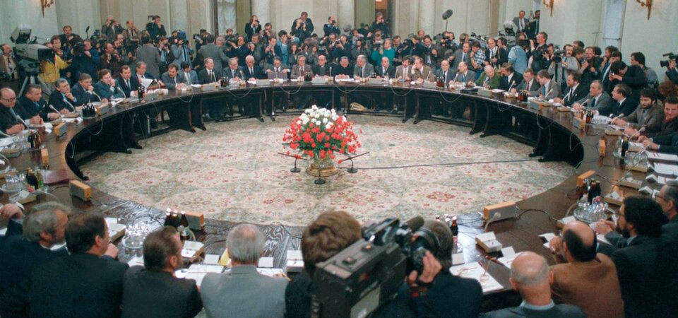 Zdjęcie przedstawia okrągły, drewniany stół. Wokół niego na krzesłach siedzą ludzie. Na środku znajduje się bukiet biało‑czerwonych kwiatów. Na stole znajdują się dokumenty, szklanki oraz mikrofony. Pod ścianą stoją dziennikarze z kamerami i aparatami fotograficznymi.