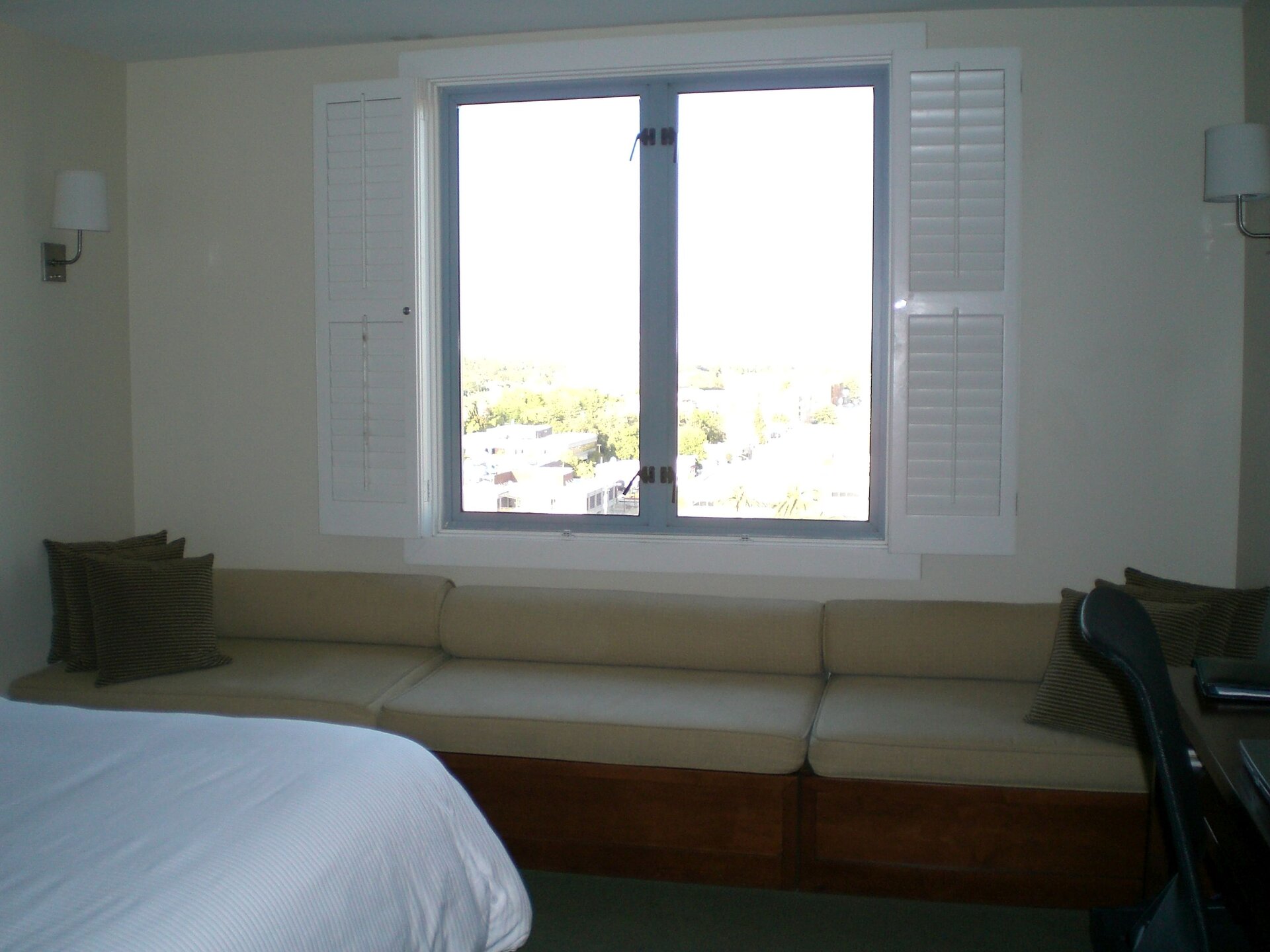 Ilustracja przedstawia wnętrze pokoju z oknem. Pod ścianą stoi długa kanapa. Nad nią znajduje się okno z białymi okiennicami. Przez nie do pomieszczenia wpada dzienne światło.