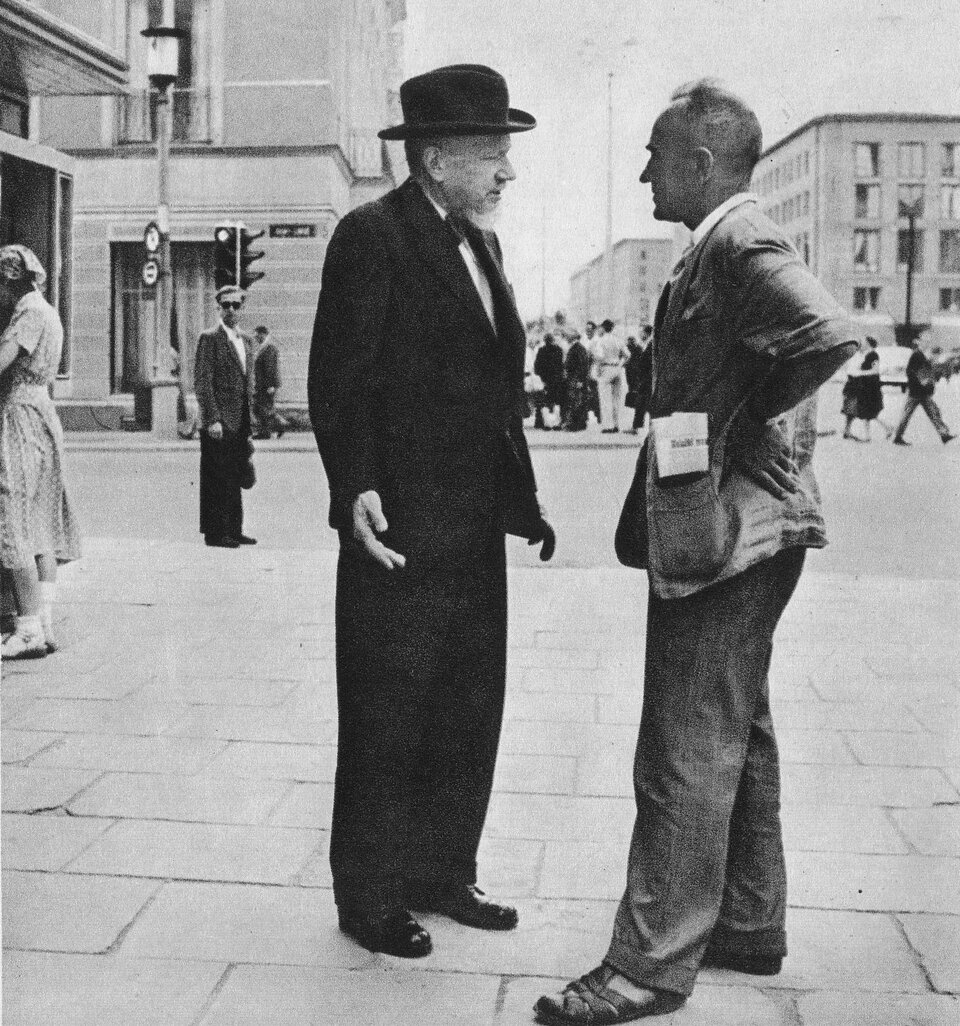 Zdjęcie przedstawia dwóch mężczyzn stojących na chodniku w dużym mieście. Postać stojąca po lewej stronie jest w średnim wieku, ukazuje prawy profil. Ma zwężone oczy i prosty nos. Jego broda jest starannie ostrzyżona, a wąsy niewielkie. Na głowie ma kapelusz. Jest ubrany w garnitur. Po prawej stronie stoi dojrzały mężczyzna. Ukazuje lewy profil. Łysieje i ma przerzedzone włosy. Jego brwi są krzaczaste, a nos długi. Ma gładko ogolone policzki i podbródek. Jest ubrany w jasny garnitur. Z kieszeni marynarki wystaje złożona gazeta. W tle skrzyżowane ulice i kilkukondygnacyjne kamienice i liczni przechodnie.