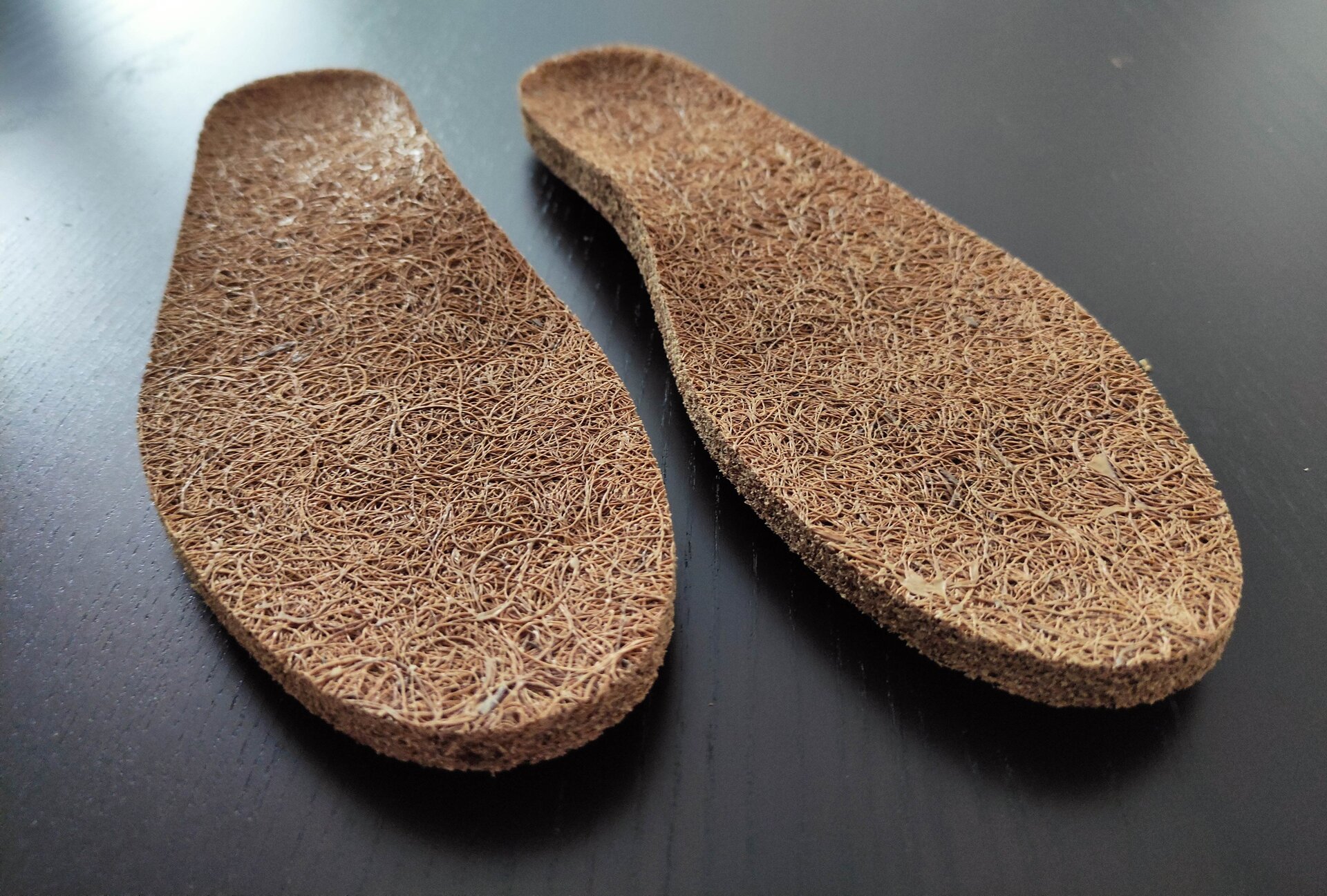Zdjęcie przedstawia dwie płaskie, cienkie wkładki w kształcie stopy wykonane z włókna kokosowego.