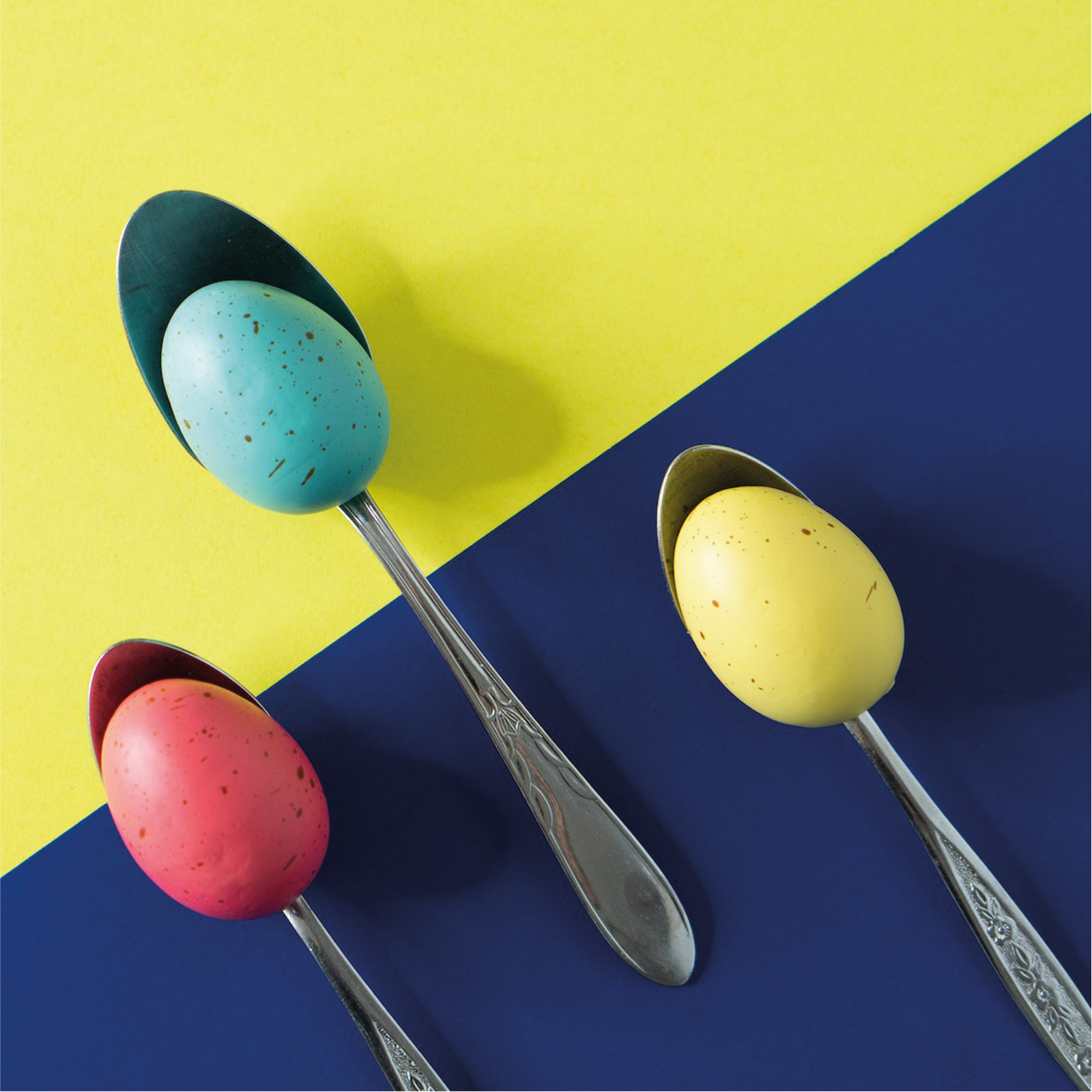 Ilustracja przedstawia żółto‑niebieski blat, na którym leżą trzy łyżki, a na nich jajka w kolorach czerwonym, żółtym i niebieskim.