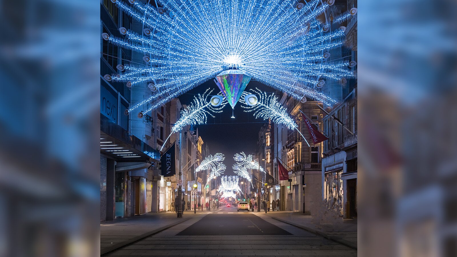 Zdjęcie przedstawia niewielką uliczkę udekorowaną ozdobnym oświetleniem. Iluminacja została wykonana na kształt rozłożonego pawiego ogona z kryształem pośrodku łączących się nad środkiem ulicy dwóch pawich piór. Dekoracje świecą się zimnym, niebieskawo‑białym światłem. Wzdłuż uliczki widoczne są kamienice, z witrynami sklepowymi na poziomie chodnika. 