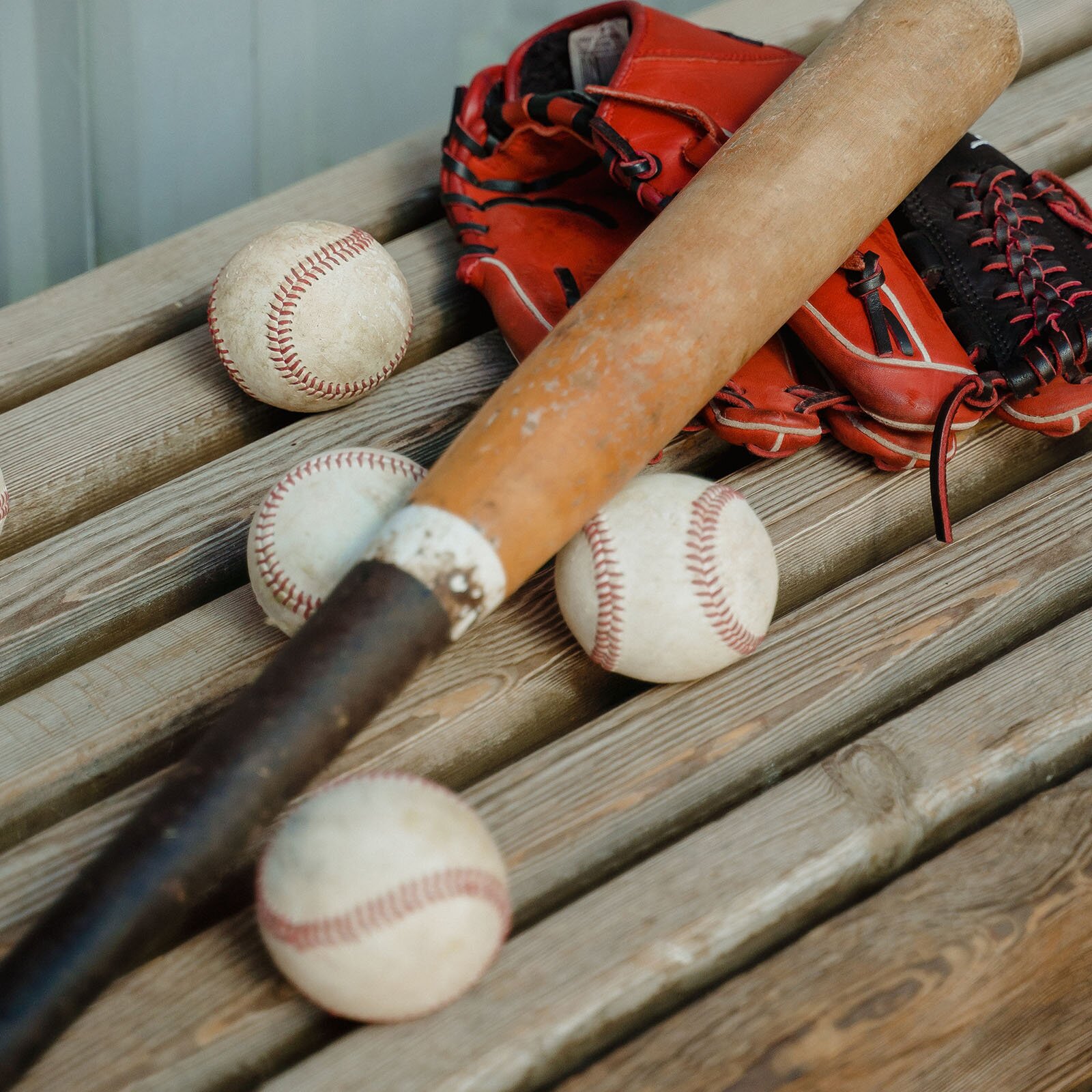 Ilustracja przedstawia drewnianą ławkę, na której leżą cztery piłki do gry w  Baseball czerwono‑czarna rękawica do łapania piłek oraz drewniany kij do tej samej gry.