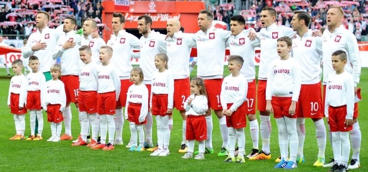 Zdjęcie przedstawia reprezentację Polski wraz z wprowadzającymi piłkarzy na boisko dziećmi. Piłkarze  i dzieci ubrani są w biało- czerwone stroje, białe koszulki, czerwone spodenki, białe długie skarpetki. Na koszulkach dzieci nazwa sponsora, na koszulkach piłkarzy godło z orłem. Piłkarze trzymają się za ramiona. Wszyscy śpiewają hymn  przed meczem.