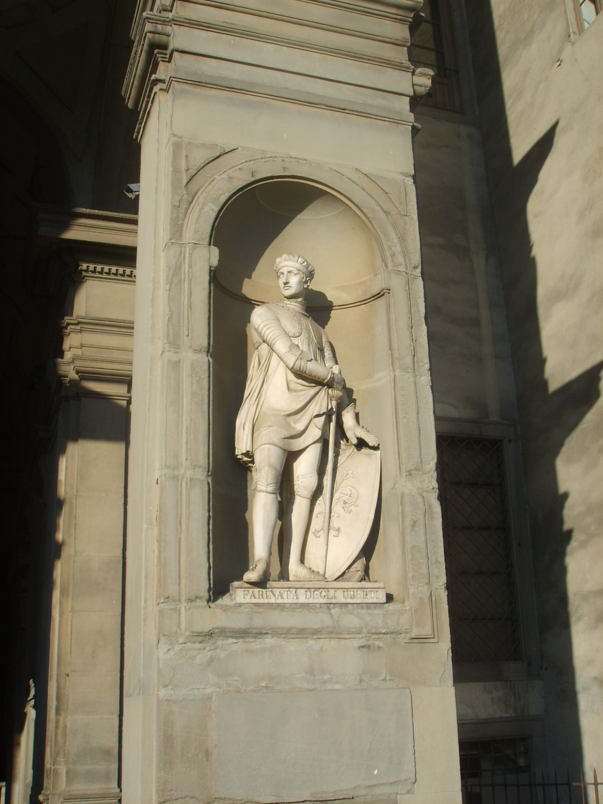 Fotografia przedstawia posąg młodego mężczyzny ubranego w zbroję zakrywającą klatkę piersiową oraz zwiewny materiał okrywający tułów. Na kolanach ma nakolanniki, na dłoniach rękawice, a na nogach - obuwie. Mężczyzna ma krótkie włosy oraz opaskę na czole. W jednej dłoni trzyma miecz, a w drugiej tarczę. Rzeźba mężczyzny została umieszczona w niszy, w ścianie budynku.