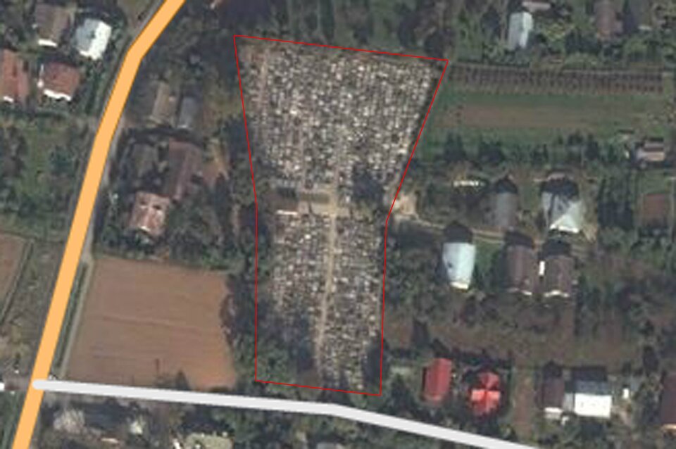 Fotografia cmentarza wykonana z lotu ptaka. Teren cmentarza zaznaczony czerwoną linią w kształcie zbliżonym do prostokąta. Obok budynki, pola i drogi. 