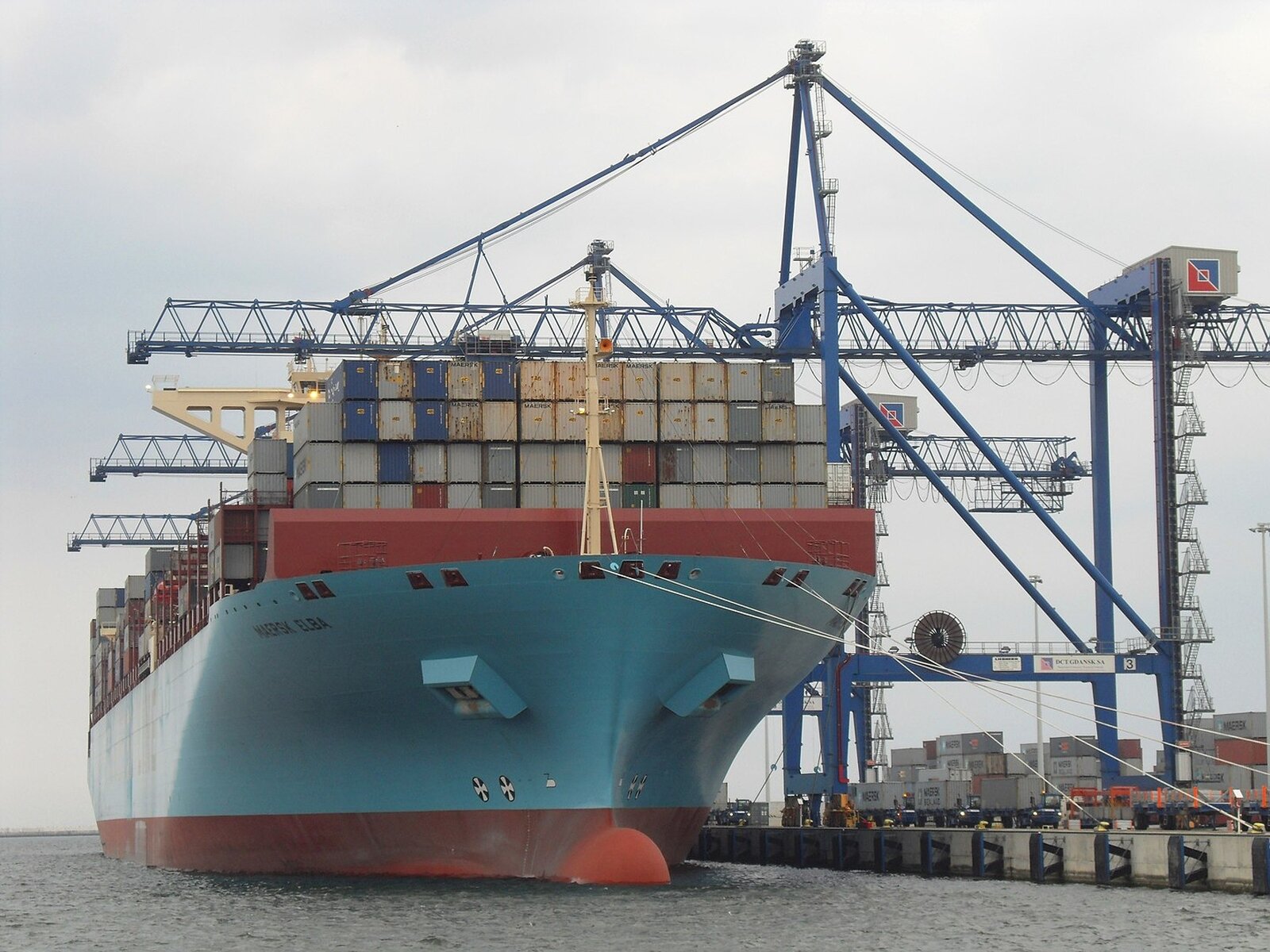 Na zdjęciu bardzo duży statek z setkami umieszczonych na nim jeden na drugim kontenerów. Po prawej stronie żurawie, za pomocą których odbywa się załadunek towaru.