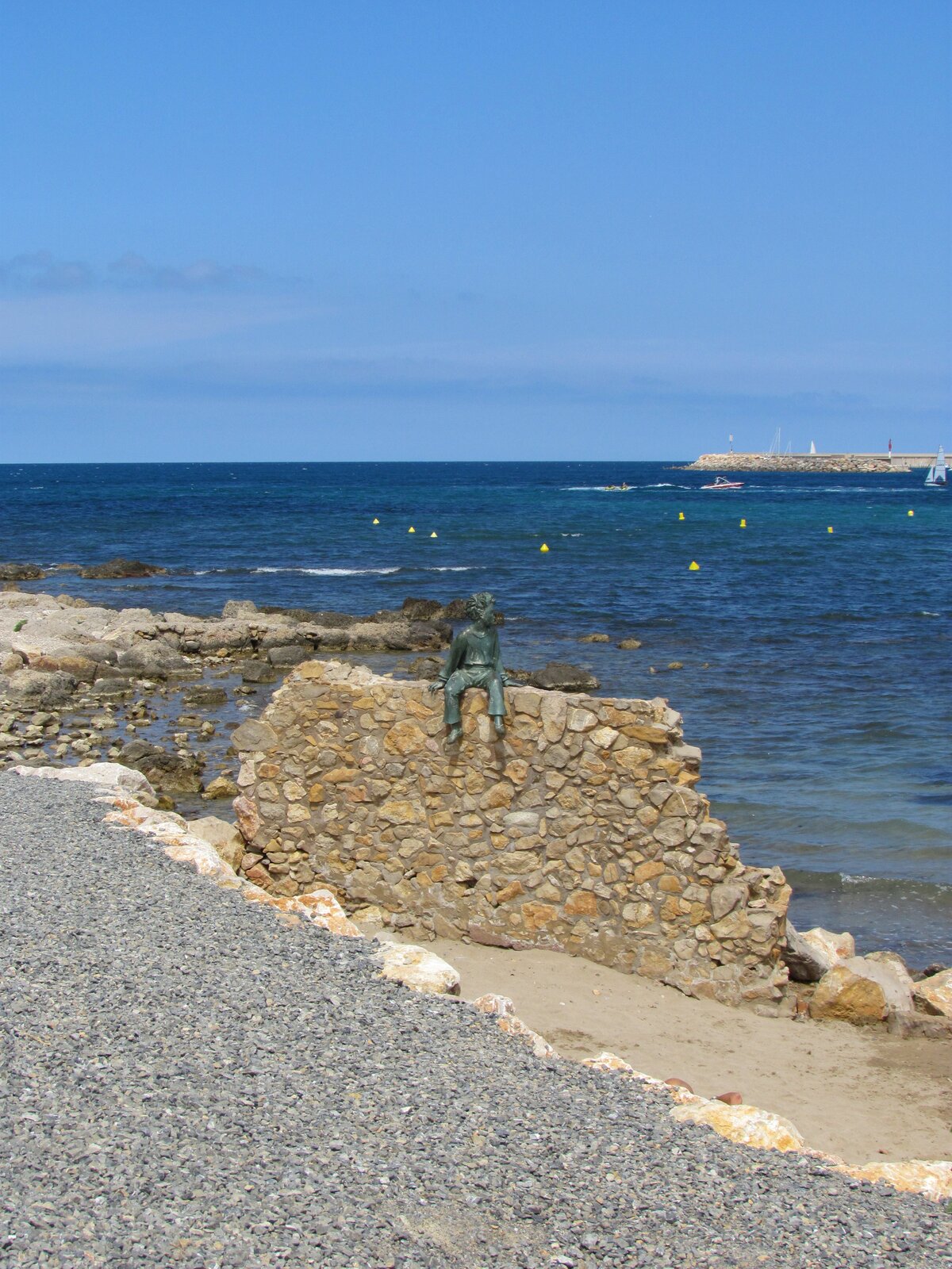 Na zdjęciu widoczna jest umiejscowiona w centrum rzeźba Małego Księcia, która siedzi na kamiennym murze i patrzy w prawo. W lewym dolnym rogu widoczny jest kamienisty brzeg. W tle morze i niebieskie bezchmurne niebo. 