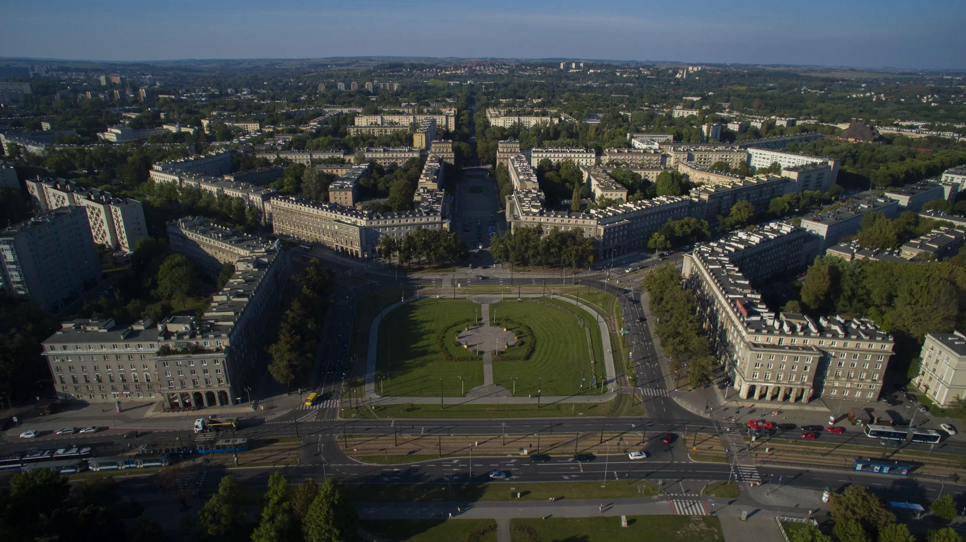 Fotografia przedstawia Nową Hutę w Krakowie, kompleks budynków mieszkalnych. W centrum ilustracji znajduje się duży obszar zieleni, trawnik, który stanowi rondo dla trzypasmowej ulicy. Od zielonego skweru, promieniście, odchodzą ulice, wzdłuż których ciągną się bloki, wytyczając granice osiedli. Zabudowa osiedli jest luźna, a wkomponowana w przestrzeń zieleń zapewnia mieszkańcom komfort życia.