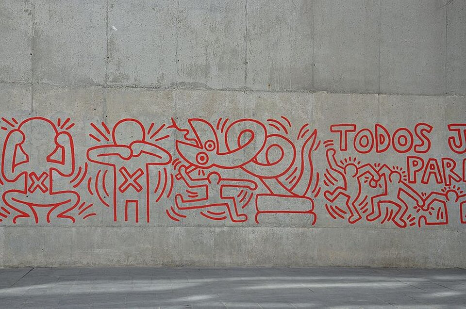 Ilustracja przedstawia dzieło Keitha Haringa: mural w Barcelonie. Na ilustracji znajduje się fragment muru. Możemy na nim dostrzec liczne rysunki wykonane linią tej samej grubości w kolorze czerwonym. Obok grafik znajdują się także podpisy wykonane czerwoną farbą. Artysta słynie z tworzenia dzieł w nurtach pop‑art oraz street art.