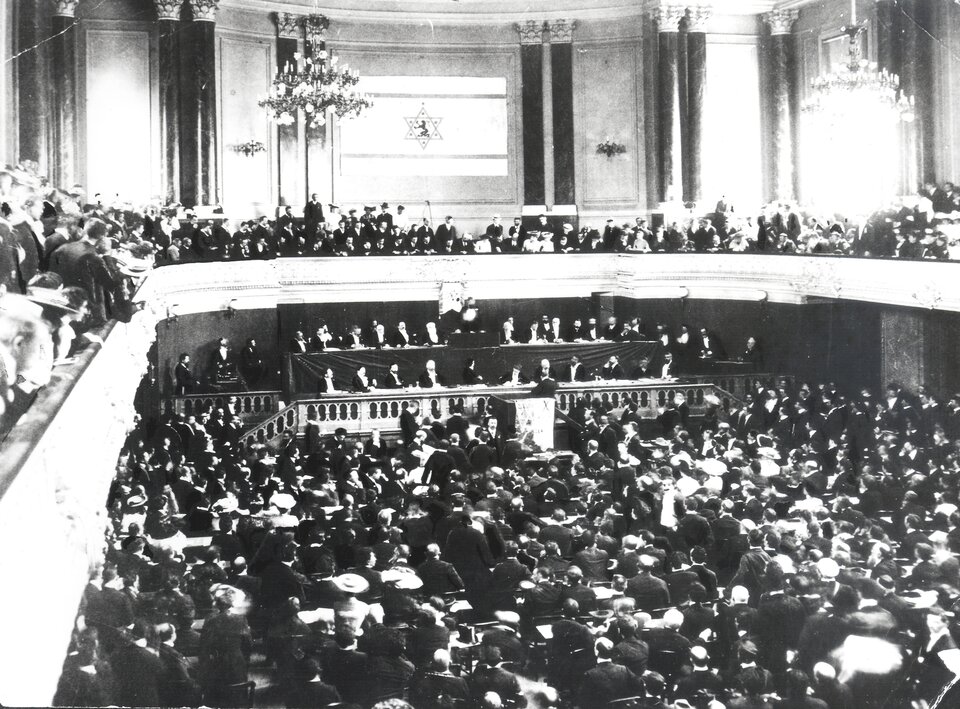 Czarno‑białe zdjęcie przedstawia dużą salę wypełnioną ludźmi. Dookoła sali znajduje się balkon również wypełniony ludźmi. W sali w przedniej części są dwa rzędy stołów, przy których siedzą najważniejsi politycy. Jest tam również mównica. Stoi przy niej mężczyzna. Z sufitu zwisają żyrandole. Na ścianie nad balkonem umieszczona jest duża flaga państwa Izrael. 