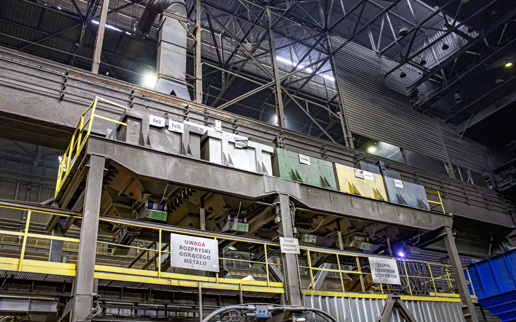 Na zdjęciu przedstawiono zasobniki dodatków stopowych. W centralnej części zdjęcia znajduje się stalowa dwupoziomowa konstrukcja. Pierwszy poziom zabezpieczony jest żółtą barierką ochronną. Drugi poziom posadowiony jest na sześciu pionowych stalowych filarach. Na filarach osadzona jest galeria na której widać pięć kolorowych zasobników. Zasobniki są o kształcie zbliżonym do prostopadłościanuW tle zdjęcia widać dobrze oświetlone wnętrze dużego obiektu przemysłowego.