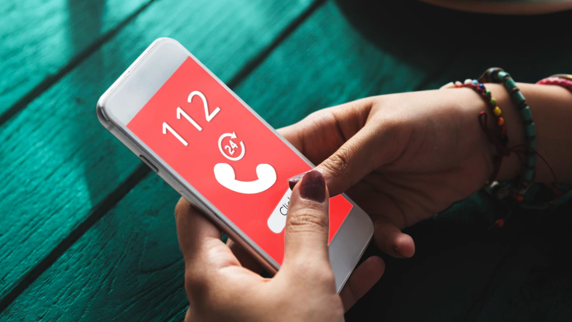 Zdjęcie przedstawia zbliżenie na telefon komórkowy, który znajduje się w kobiecych dłoniach. Na ekranie telefonu wyświetla się numer 112, obok znajduje się symbol słuchawki oraz znaczek z liczbą 24. Poniżej znajduje się przycisk do nawiązania połączenia. Na nadgarstku kobieta ma założone bransoletki oraz ma pomalowane paznokcie.