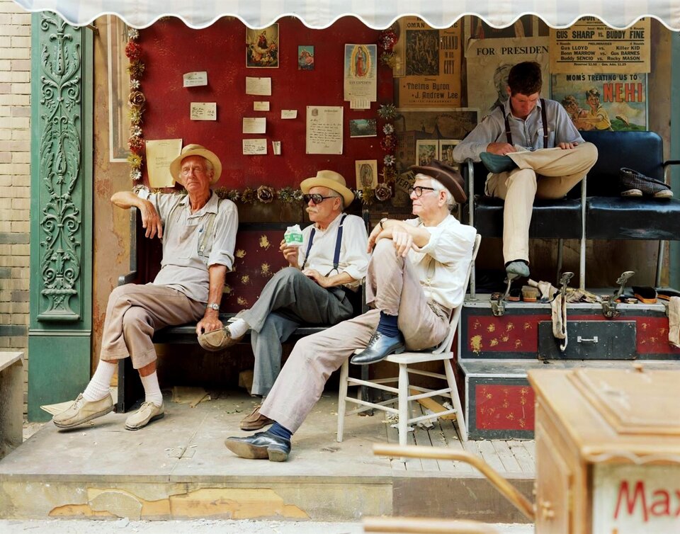 Ilustracja przedstawia fotografię barwną na której znajdują się cztery osoby. Na planie pierwszym siedzą trzej mężczyźni na krzesłach przed budynkiem, którego fragment znajduje się za ich plecami. Mężczyźni są w kapeluszach i patrzą w tym samym kierunku. Wydają się być w podobnym wieku. Czwarty mężczyzna jest młody, siedzi swobodnie na krześle, nieco z tyłu. Ma głowę pochyloną i wydaje się być czymś zajęty. 