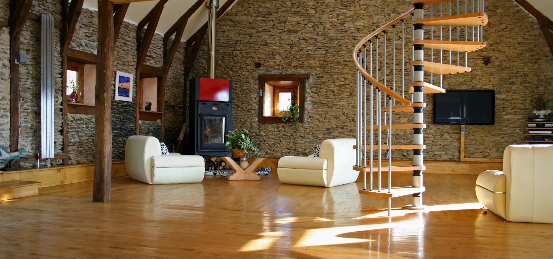 Ilustracja przedstawia pokój, ze ścianami pokrytymi dekoracyjnymi kamieniami. Ramy okienne, listwy podłogowe, podpory, podpory dachowe oraz schody wykonane są z drewna. Po lewej stronie pokoju widać podłużny 5 żebrowy grzejnik, dwa okna, a pomiędzy nimi niebieski obraz w  białej ramie. W lewym rogu pokoju znajduje się czarny piec, z czerwoną blachą nad szklanymi drzwiczkami. Pod piecem leży drewno kominkowe. Przed piecem znajdują się dwa białe fotele z biało czarnymi poduszkami, a pomiędzy nimi znajduje się drewniany stół ze szklanym blatem. Na stole znajduje się roślina w koszyku. Po prawej stronie pieca znajduje się kolejne drewniane okno z rośliną na parapecie. Po prawej stronie foteli są drewniane kręcone schody z białymi podpórkami.  W prawej części pokoju widać płaski wyłączony telewizor powieszony na ścianie. Po jego prawej stronie stoi półka z dokumentami, a naprzeciwko kolejny biały fotel.