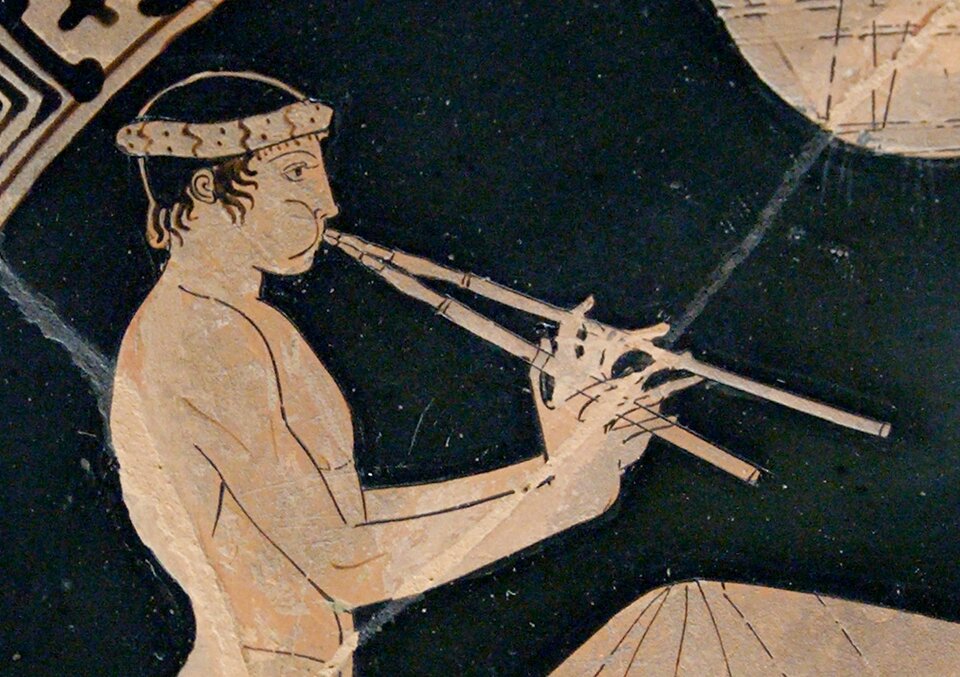 Fragment czerwonofigurowego malowidła na naczyniu przedstawia siedzącego mężczyznę grającego na aulosie, starożytnym instrumencie dętym złożonym z dwóch piszczałek. Muzyk trzyma obie piszczałki w dłoniach i przykłada je do ust. Ma na głowie opaskę, a jego jasna sylwetka kontrastuje z czarnym tłem.