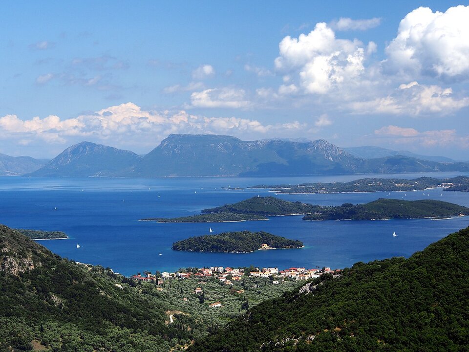 Ilustracja przedstawia Wyspy Jońskie w Grecji. Na pierwszym planie są pagórki porośnięte zielona roślinnością, dalej niebieska tafla wody, na niej kilka wysepek, w oddali widoczna większa górzysta wyspa porośnięta lasem. Po po lewej stronie jest południowy kraniec wyspy Sparta.
