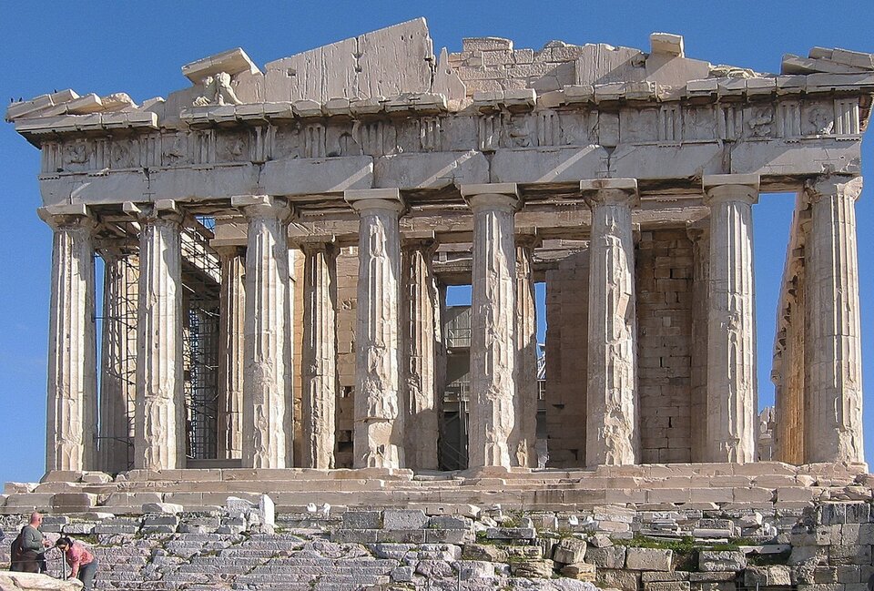 Na fotografii znajdują się ruiny Partenonu. Jest to marmurowa budowla z licznymi kolumnami. W jej lewej części widoczne są metalowe rusztowania. 