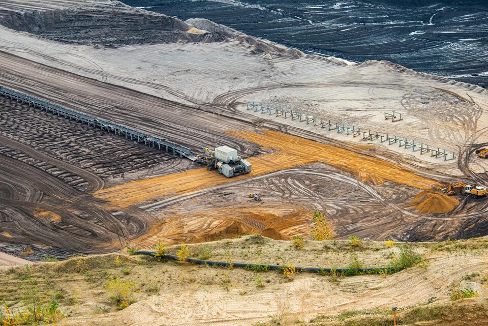 Krajobraz odkrywkowej kopalni surowców mineralnych z widocznymi maszynami górniczymi, wyraźnymi śladami eksploatacji terenu oraz pasami przetworzonej ziemi i piasku.