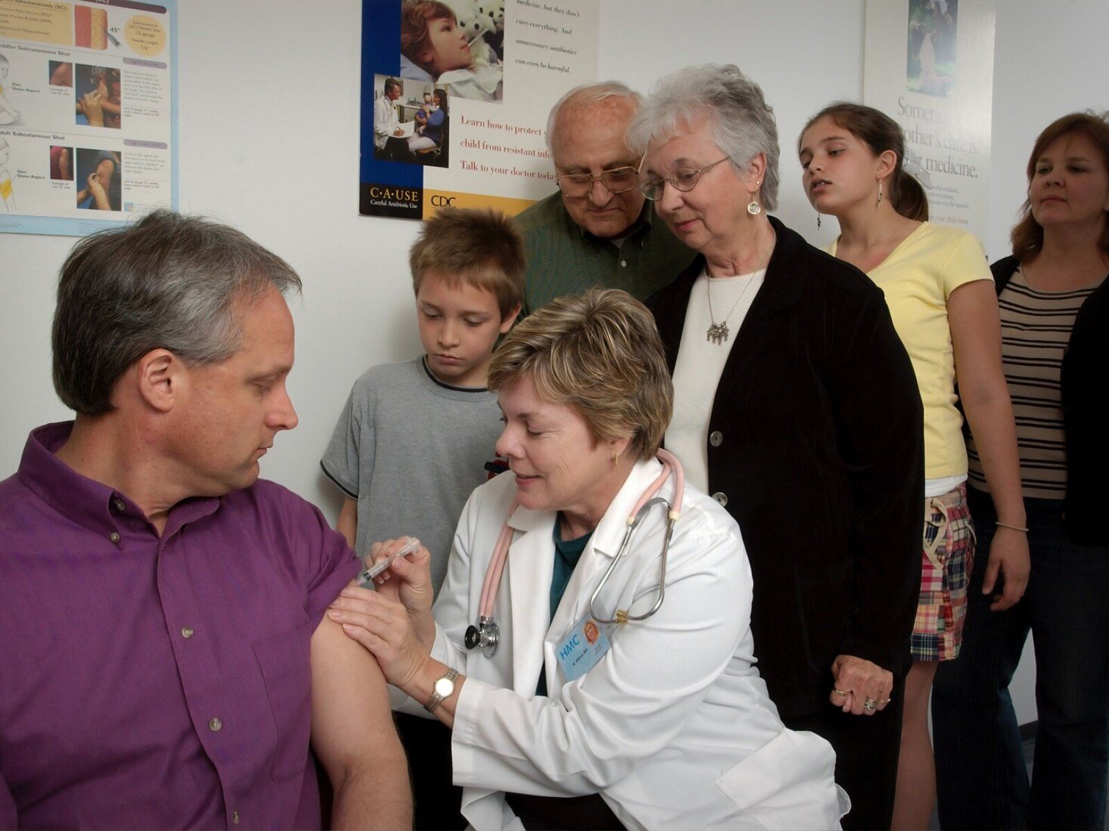 Zdjęcie przedstawia pielęgniarkę, ubraną w biały fartuch i ze stetoskopem na szyi, robiącą zastrzyk w ramię mężczyźnie w średnim wieku. Za nimi inni pacjenci stojący w kolejce.