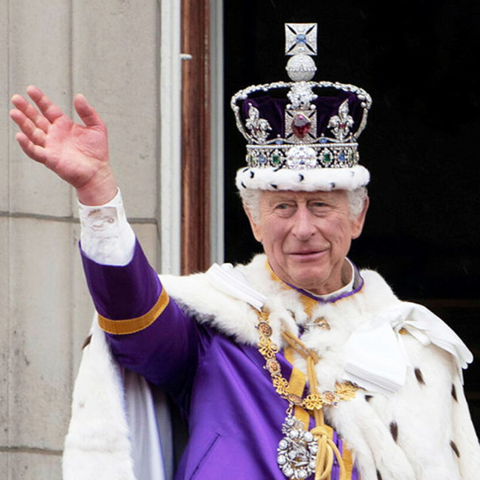 Zdjęcie przedstawia króla Karola III, w ujęciu od pasa w górę. Widoczny jest mężczyzna w starszym wieku, ubrany we fioletową górę, na którą ma narzucony gronostajowy płaszcz, a na piersi zawieszone złote insygnia królewskie. Na głowie ma założoną koronę. Jest zwrócony lekko w lewo, patrzy przed siebie, a prawą ręką uniesioną nad głowę wykonuje gest pozdrowienia.