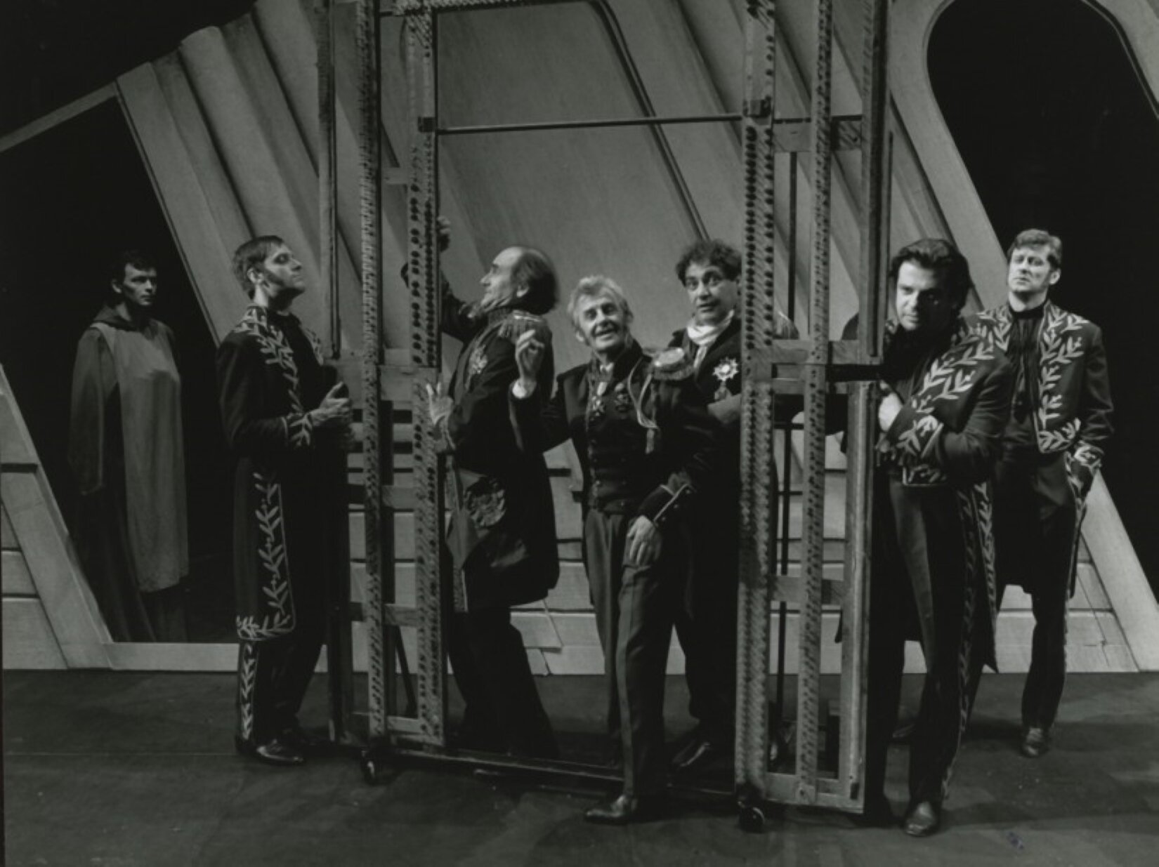 Ilustracja przedstawia fotografię Wojciecha Plewińskiego. Znajduje się na niej scena z „Dziadów - dwanaście improwizacji według Mickiewicza” w reżyserii Jerzego Grzegorzewskiego w dniu 24.06.1995 roku w Starym Teatrze w Krakowie. Na fotografii jest 7 mężczyzn. Na pierwszym planie stoi wysoka rama na małych kółkach. Przechodzi przez nią jeden z mężczyzn. Ma na sobie dwurzędowy żakiet z ozdobnymi naramiennikami, długie spodnie z kantem i buty. Jego prawa ręka jest uniesiona w górę. Za nim stoi dwóch mężczyzn. Postać z lewej strony stoi bokiem i trzyma ramę. Ma na sobie długą marynarkę z ozdobnymi naramiennikami i długie spodnie. Postać z prawej strony ma biały szal pod szyją i marynarkę z przypiętym orderem. Po lewej stronie ramy, bokiem stoi mężczyzna ubrany w ozdobny frak. Obie dłonie trzyma na ramie. Po prawej stronie ramy stoi dwóch mężczyzn ubranych w takie same ozdobne fraki. Jeden z nich opiera się o ramę, a drugi trzyma ręce w kieszeniach. W tle znajduje się ściana z dwoma otworami. W otworze z lewej strony stoi mężczyzna w długim, luźnym ubraniu. Fotografia jest w kolorystyce czarno‑białej.  