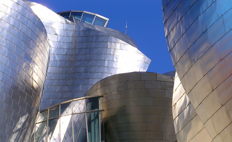 Fotografia przedstawia budynek na tle intensywnie niebieskiego nieba. Z lewej strony, z przodu, a także z prawej strony, widoczne są ściany srebrnego budynku, którego powierzchnia odbija światło słoneczne. Składają się one z wielu srebrnych, prostokątnych lśniących elementów, sprawiających, że budynek przypomina powierzchnię folii aluminiowej. Ściany są zaokrąglone, pozbawione linii prostych, występujących tylko w krawędziach prostokątnych cząstek. Po lewej stronie, u dołu fotografii, widać oszklony fragment budynku, składający się z dwóch rzędów – górnych kwadratów, oddzielonych od podłużnych prostokątów na dole. Widać w nich odbicie budynku. 