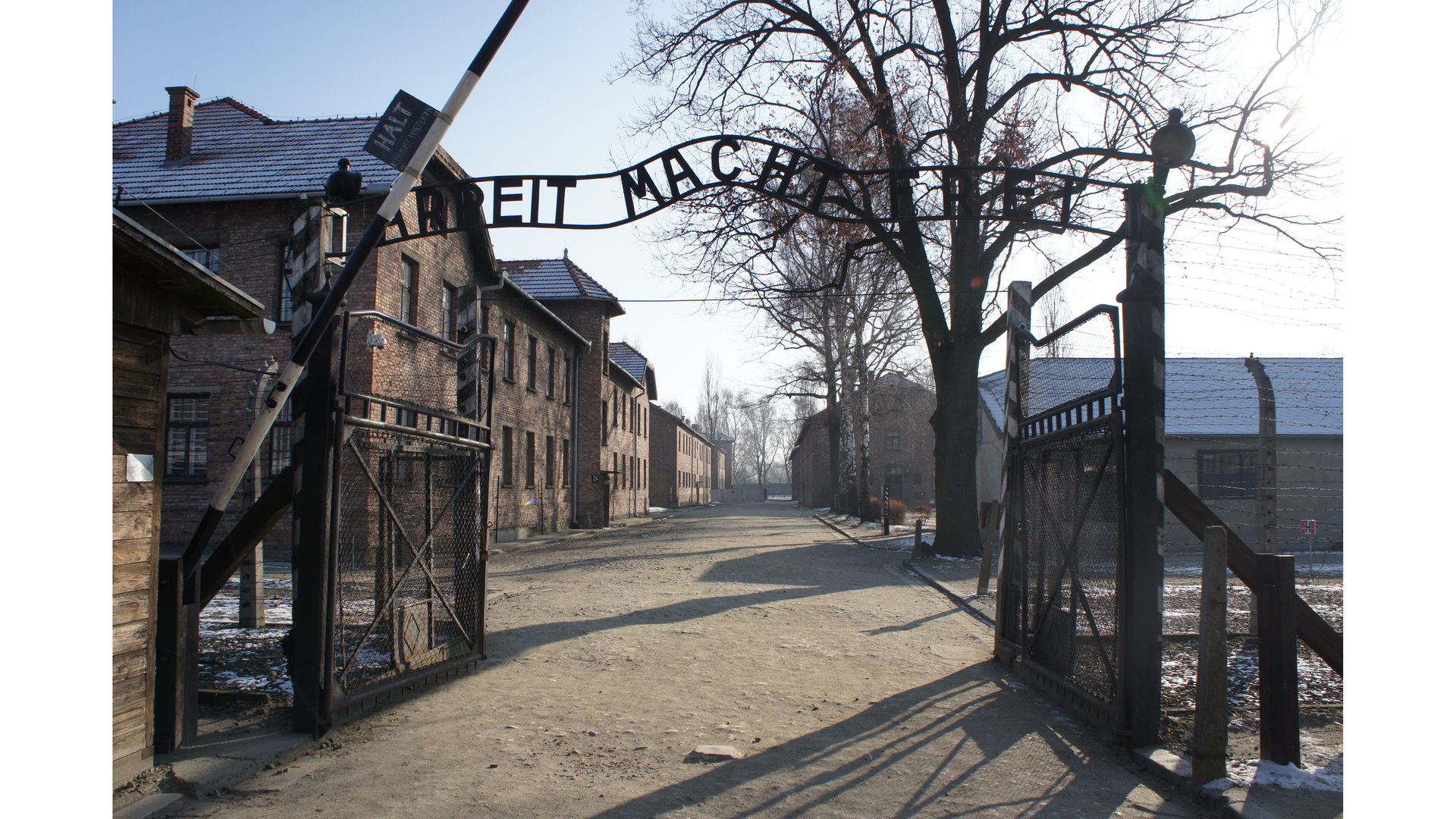 Na zdjęciu widzimy bramę wjazdową, przed nią podniesiony szlaban. W dalszej części niskie ceglane zabudowania.