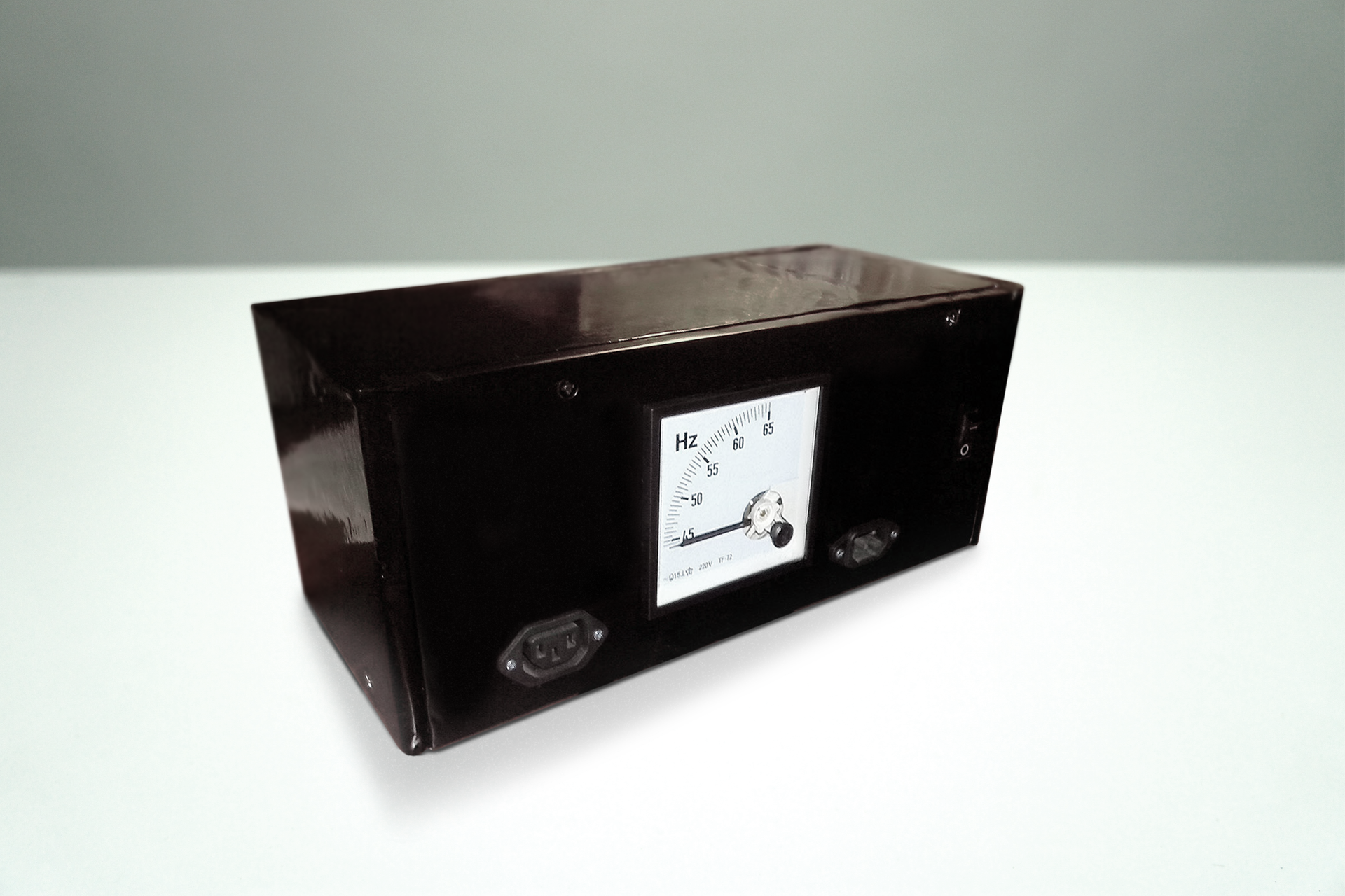 Zdjęcie przedstawia częstościomierz stojący na jasnym stole. Tło jasne. Częstościomierz ma on postać czarnej skrzynki - prostopadłościanu z analogowym wskaźnikiem na tle białego kwadratowego pola ze skalą w kształcie łuku.