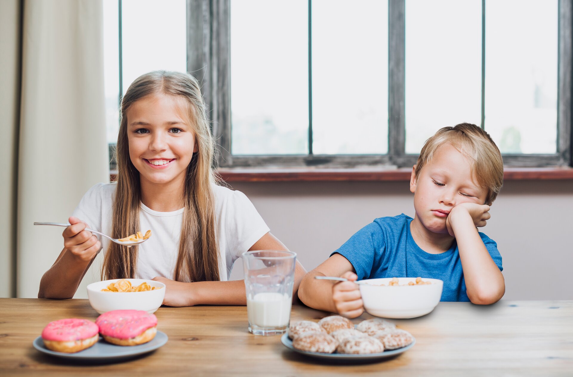 Zdjęcie przedstawia dziewczynkę i małego chłopca, którzy siedzą przy stole i jedzą posiłek. Po lewej stronie widoczna jest uśmiechnięta dziewczynka z długimi, jasnymi włosami, ubrana w białą bluzkę z krótkim rękawem. Dziewczynka w podniesionej prawej ręce trzyma łyżkę, na której widoczne są płatki kukurydziane. Przed nią na blacie stoi biała miseczka z płatkami oraz niebieski talerz z dwoma pączkami polanymi różowym lukrem. Na prawo od niej widoczny jest mały chłopiec z krótkimi, jasnymi włosami, ubrany w niebieską bluzkę z krótkim rękawem. Chłopiec ma zamknięte oczy, lewą ręką podpiera opadającą na bok głowę, a w prawej ręce leżącej na blacie trzyma łyżkę zanurzoną w misce. Przed nim na blacie stoi biała miska z płatkami oraz niebieski talerz z ciastkami i szklanka z mlekiem. W tle widoczne jest duże okno.  
