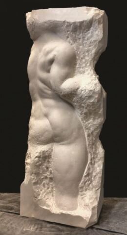 Zdjęcie przedstawia rzeźbę w stylu non finito. W podłużnym, wysokim, jasnym kamieniu wykuto ciało mężczyzny, przedstawionego tyłem. Sylwetka mężczyzny jest wysportowana, z widocznymi mięśniami. Artysta dokończył jedynie plecy, część prawej ręki, pośladki oraz część prawej nogi. Reszta rzeźby ma nieregularny kształt oraz nierówną powierzchnię z śladami po dłucie. W dolnej części rzeźby marmur pozostał gładki i nietknięty nawet przez artystę. Rzeźba stoi na drewnianym podłożu, na czarnym tle.