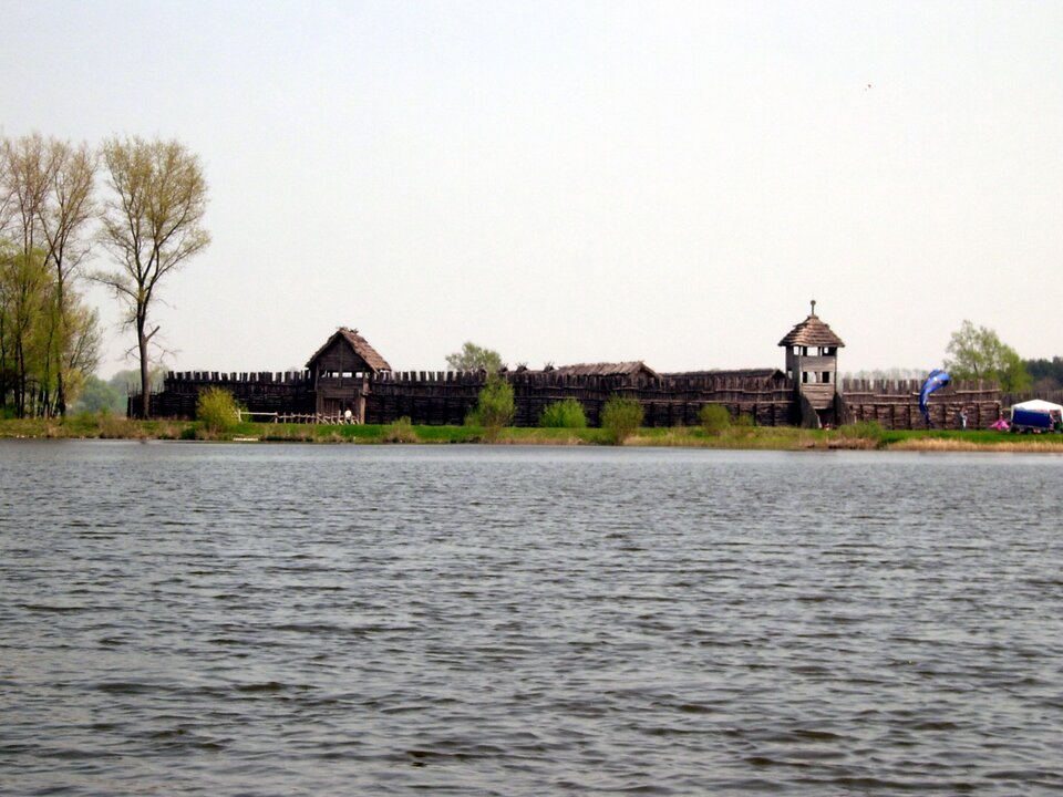 Fotografia przedstawia zrekonstruowaną część obronnej osady kultury łużyckiej w Biskupinie, widzianą od strony jeziora. Na pierwszym planie znajduje się powierzchnia jeziora. W oddali rozciąga się  widok na drewnianą palisadę, zbudowaną z pionowo ustawionych, zaostrzonych na końcach bali. Do osady prowadzi brama, a nad ogrodzeniem wystają dachy chat z dwuspadowymi strzechami. Sceneria otoczona jest roślinnością – w tle widoczna jest linia drzew porastających brzeg jeziora.