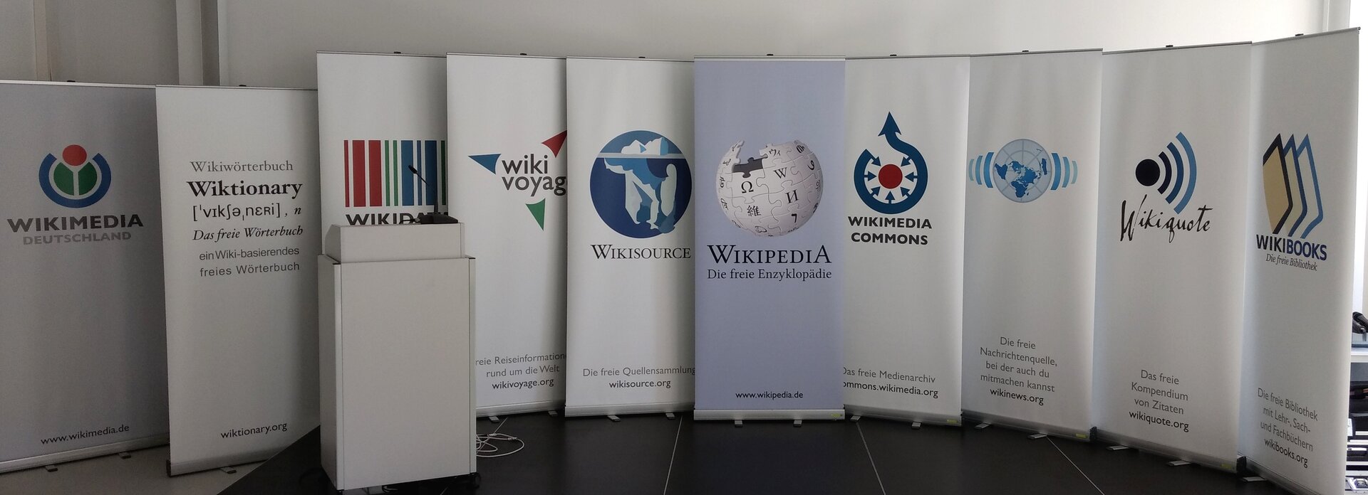 Fotografia przedstawiająca plakaty typu Roll‑up należące do Wikimedia. Na scenie znajduje się 10 plakatów, przed nimi stoi mównica z mikrofonem. Od lewej przedstawiają nazwy z logo: Wikimedia Niemcy, Wikitionary, Wikidata, Wiki Voyag, Wikisource, Wikipedia Darmowa encyklopedia, Wikimedia Commons, Wiki News, Wikigoute, Wikibooks.